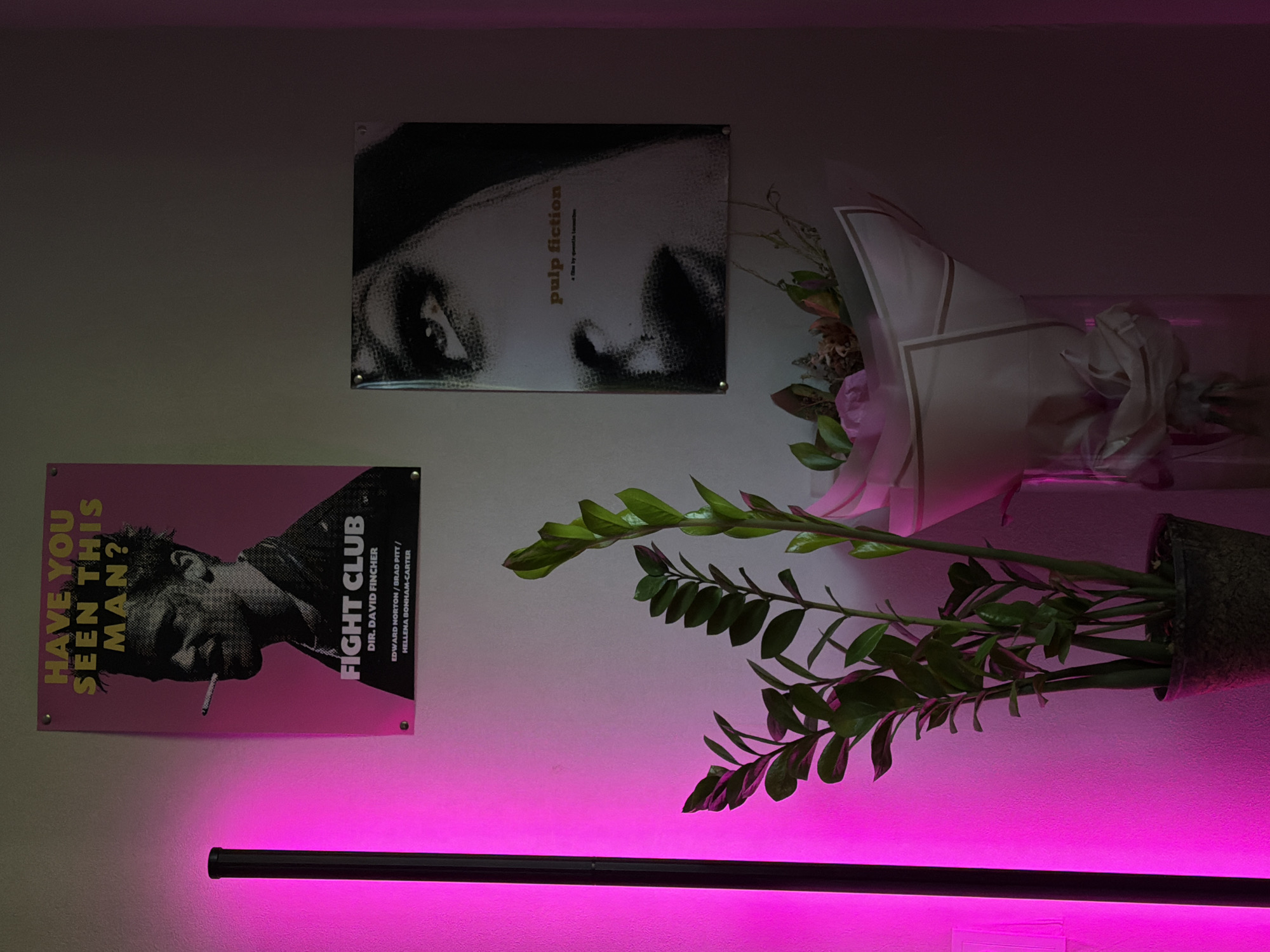 A vertical, rectangular photograph depicts an indoor scene. On the upper left, a pink poster with the text "HAVE YOU SEEN THIS MAN?" and a black and white image of Brad Pitt smoking a cigarette is attached to a light gray wall. Below this poster is a tall, black cylindrical lamp emitting a bright pink light. To the right of the Fight Club poster, a smaller, rectangular poster with a black and white close-up of a woman's eye and lips, featuring the text "pulp fiction," is also attached to the wall. In the lower left portion of the image, a potted plant with dark green leaves is visible. In the lower center, a bouquet of flowers wrapped in white paper with gold trim is placed in a vase. The overall lighting is dim, with the pink lamp providing the primary illumination on the left side of the frame.