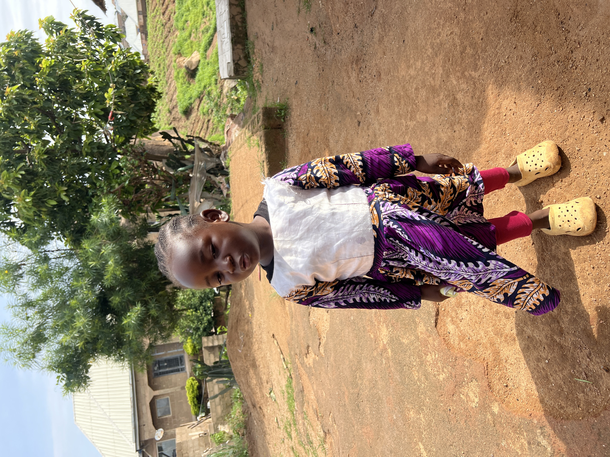 A young African child stands outdoors in a rural or semi-rural setting. The child, with braided hair and dark skin, is wearing a white top with a colorful, patterned long-sleeved dress over pink leggings. The dress features purple, black, and orange leaf-like patterns. The child is also wearing light-yellow, perforated clogs. The child is looking downwards and to the left, with their mouth slightly open.
The background shows a reddish-brown dirt ground with some scattered dry grass. Behind the child, there is a partially constructed building made of light brown bricks with a white corrugated metal roof. A large green tree with dense foliage stands to the right of the building and behind the child. Further in the background, there are more buildings with corrugated metal roofs on a sloped, grassy hill.
The overall lighting suggests a bright, sunny day. The environment appears to be a residential area, possibly in a village or the outskirts of a town. Given the visual cues of architecture, vegetation, and terrain, the scene could plausibly be located in Jos, Nigeria, a city known for its hilly landscape and mixed urban and rural characteristics.
