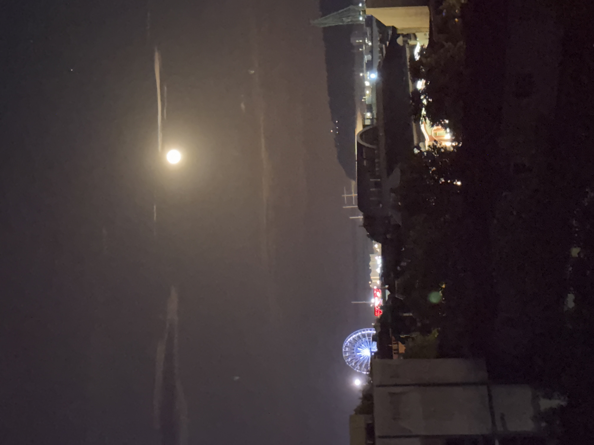 The image shows a night view of Varna, Bulgaria. In the lower part of the frame, a Ferris wheel is illuminated with blue lights, positioned near a tall, multi-story building. Above the Ferris wheel, the night sky is visible, featuring a bright moon. Further up, the cityscape includes building with illuminated windows, and a bridge structure. The right side of the image is dominated by dark trees and foliage.