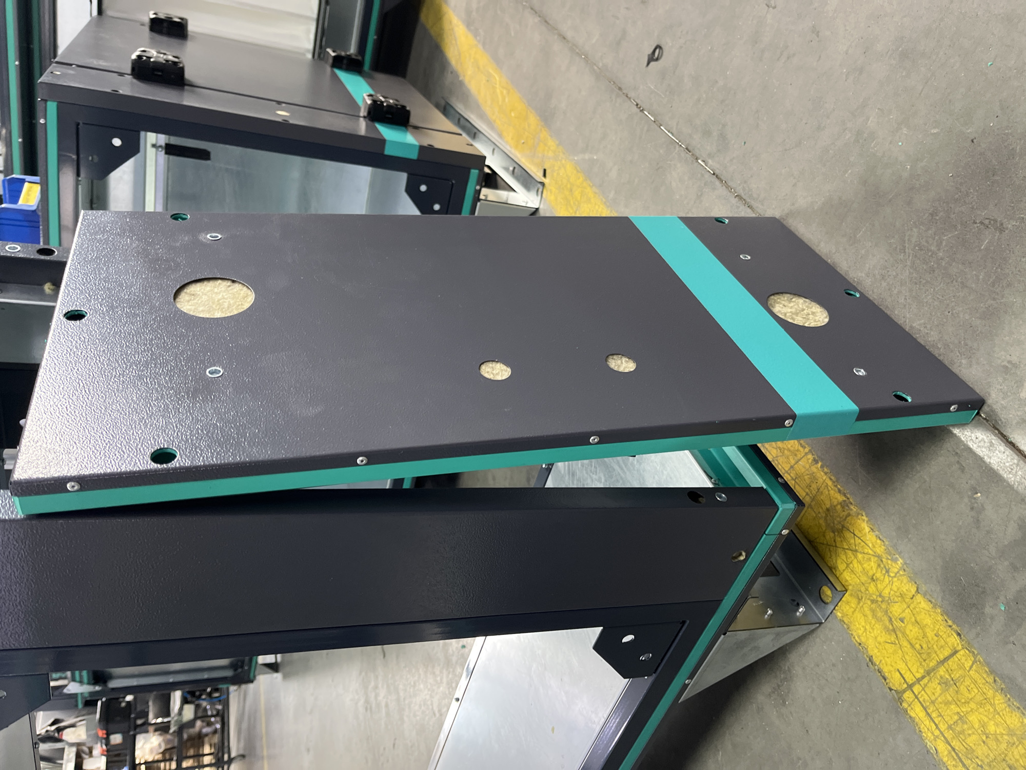 The image shows a close-up of industrial equipment, likely a ventilation unit or modular housing, in a factory or warehouse setting. The equipment is predominantly dark gray with bright teal accents. One panel of the equipment, featuring several circular cutouts with insulation material visible within, is propped open, revealing the interior. The floor is concrete, marked with a bright yellow line, suggesting a designated pathway or safety zone. In the background, other similar equipment and industrial elements are visible, including what appears to be a shopping cart and shelving units. The overall impression is one of an industrial manufacturing environment. The image does not contain any specific identifiable landmarks or details that would indicate its location in Kyiv, Ukraine.