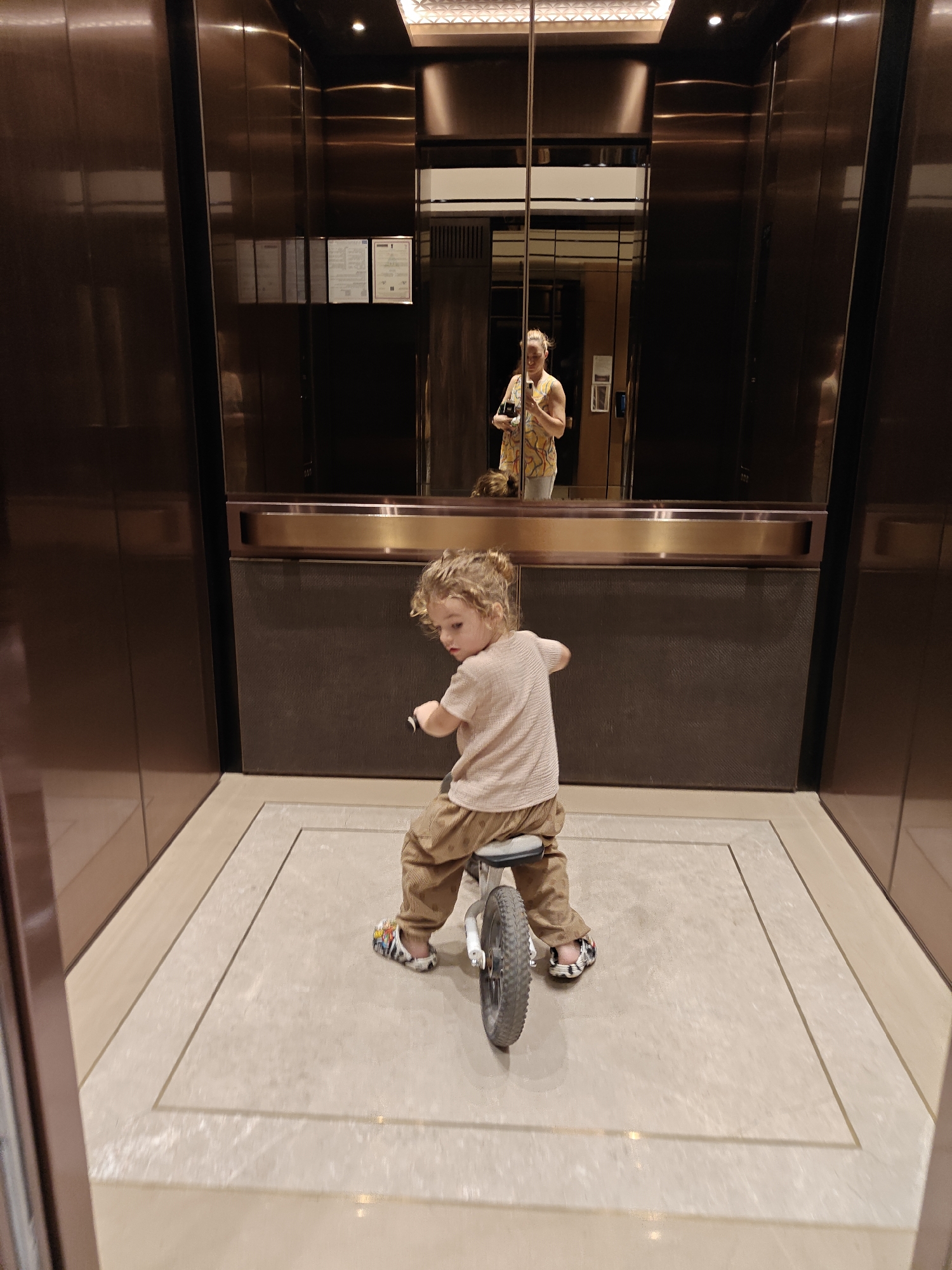 A young child with curly hair is sitting on a small bicycle in an elevator. The elevator interior features copper-colored walls and a patterned tile floor. A mirror on the back wall of the elevator reflects the person taking the photo. The location is likely Mumbai, India.