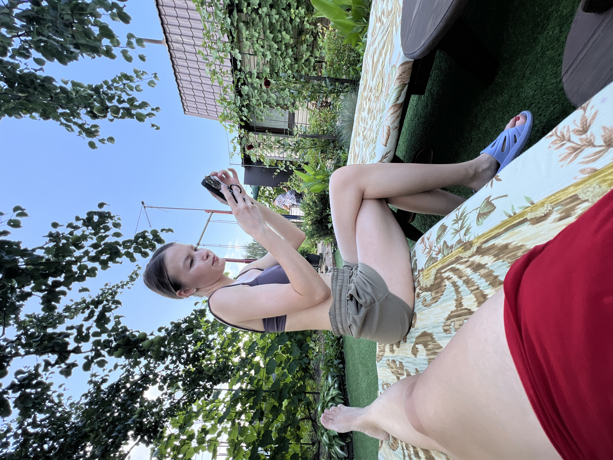 The image captures a scene in the city of Lopatino, Russia. In the foreground, a person's legs are visible, clad in red shorts, resting on a patterned lounge chair. To their right, another person, a young woman with her hair pulled back, is seated on a similar lounge chair. She is wearing a dark purple sports bra and olive-green shorts, and is engrossed in her phone, which she holds in both hands. She has blue slip-on sandals on her feet. The setting appears to be a backyard, with lush green artificial turf covering the ground. Beyond the lounge chairs, there is dense foliage from trees and vines, with a glimpse of a house with a tiled roof and a satellite dish in the background. The sky is clear and blue, suggesting a sunny day. The overall atmosphere is one of leisure and relaxation in a domestic outdoor space.