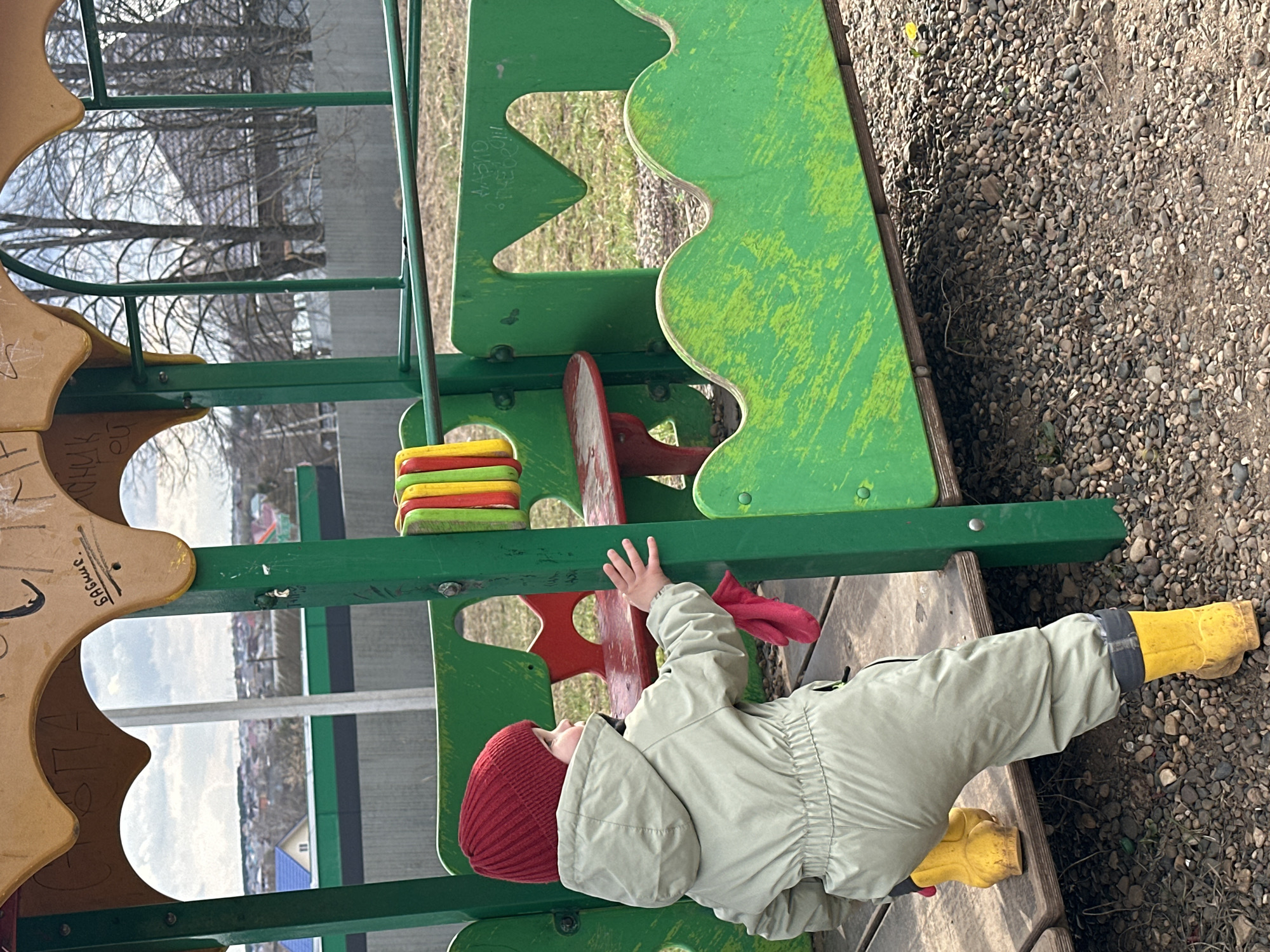 A child wearing a red beanie, light green snowsuit, and yellow boots plays on a green and yellow playground structure. The playground is surrounded by a gravel surface. In the background are trees and buildings, including a blue-roofed house. The location is potentially Ludorvay, Russia.