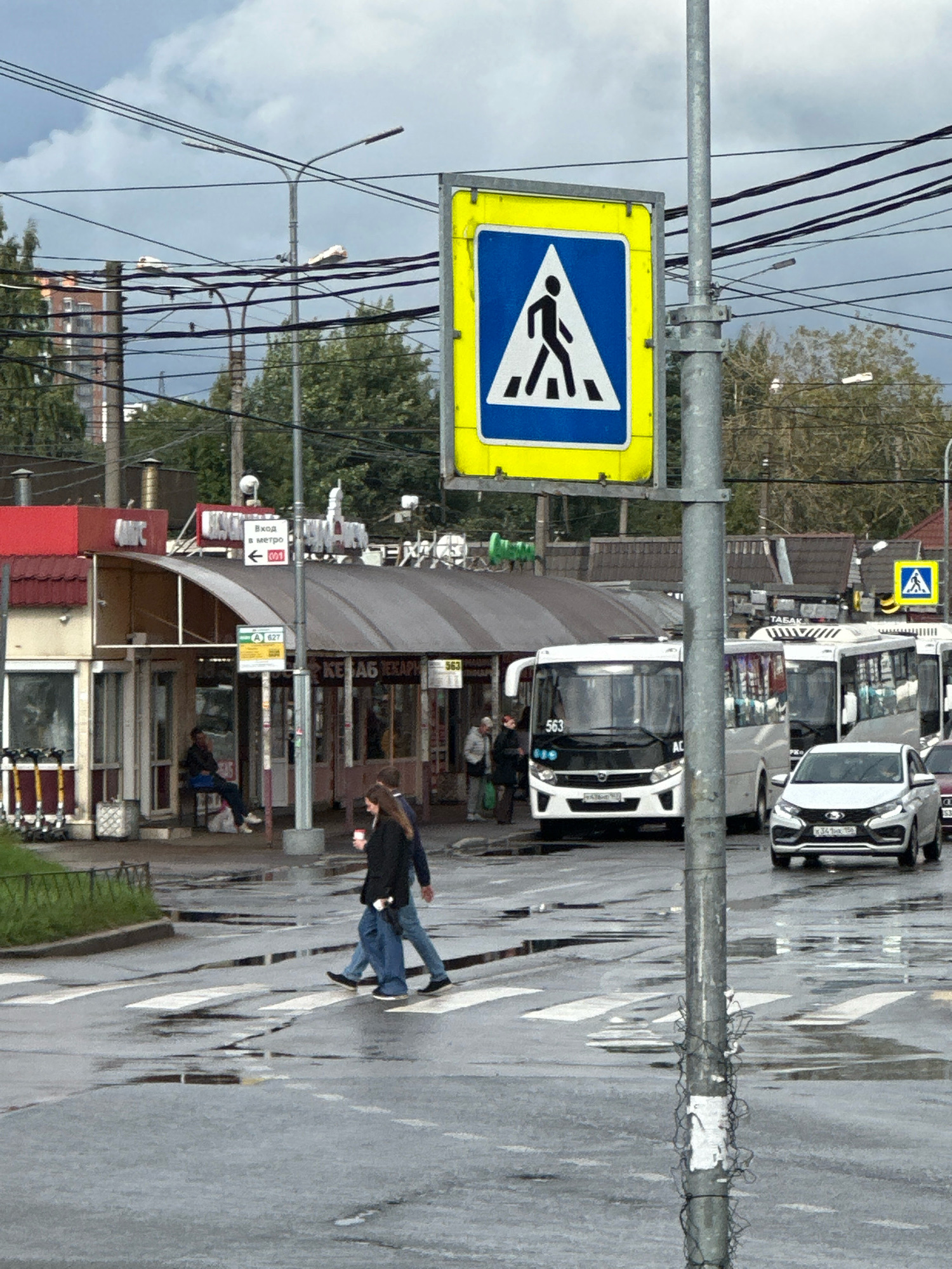 In Murino, Russia, on a cloudy day, a pedestrian crossing is active in front of a row of small shops and businesses. A prominent yellow and blue pedestrian crossing sign stands tall on a metal pole. Two individuals, a woman and a man, are captured mid-stride as they cross the wet street, with puddles reflecting the overcast sky. Behind them, several buses and a white car are stopped or moving slowly. The shops appear to be a mix of retail and food establishments, with awnings providing shelter. Overhead, a complex network of electrical wires crisscrosses the sky, adding to the urban character of the scene. The ground is wet, indicating recent rain, and the overall atmosphere suggests a typical day in a bustling Russian city.