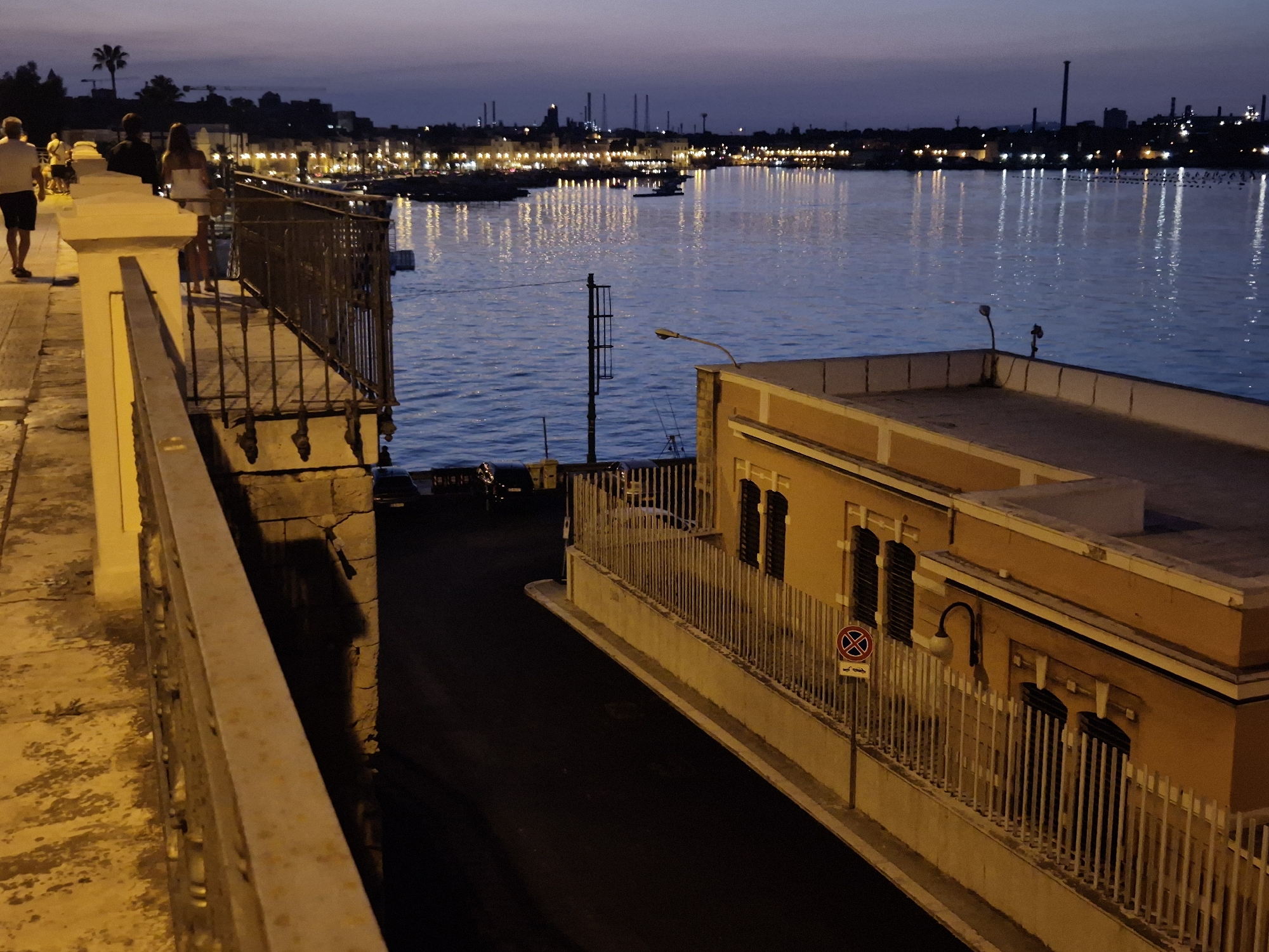 This image depicts a waterfront scene in Taranto, Italy, at dusk. The foreground features a pathway with a railing, illuminated by streetlights, casting long shadows. Several people are visible on the pathway, some walking and others standing near the railing, looking out at the water.
Beyond the pathway, the Ionian Sea is calm, reflecting the lights of the city across the water. The city skyline in the background is dotted with buildings and industrial structures, with numerous lights indicating urban activity. The sky above is a deep twilight blue, suggesting the transition from day to night.
A building with a yellow facade and a white railing is visible to the right, overlooking a dark, empty street. A "no parking" sign with a car symbol is posted on a pole in front of this building. The overall atmosphere is peaceful, with a sense of urban life continuing into the evening.