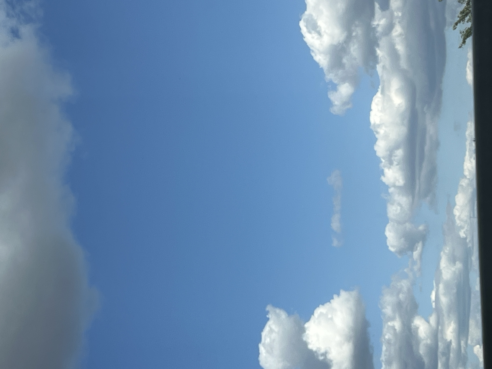 The image depicts a bright blue sky filled with scattered white and grey clouds. The lower portion of the image shows a dark, horizontal line that appears to be a barrier or the edge of a structure. The scene is devoid of any people, specific objects, or discernible actions. The provided image crops do not contain any information that would indicate a specific location in Warsaw, Poland.