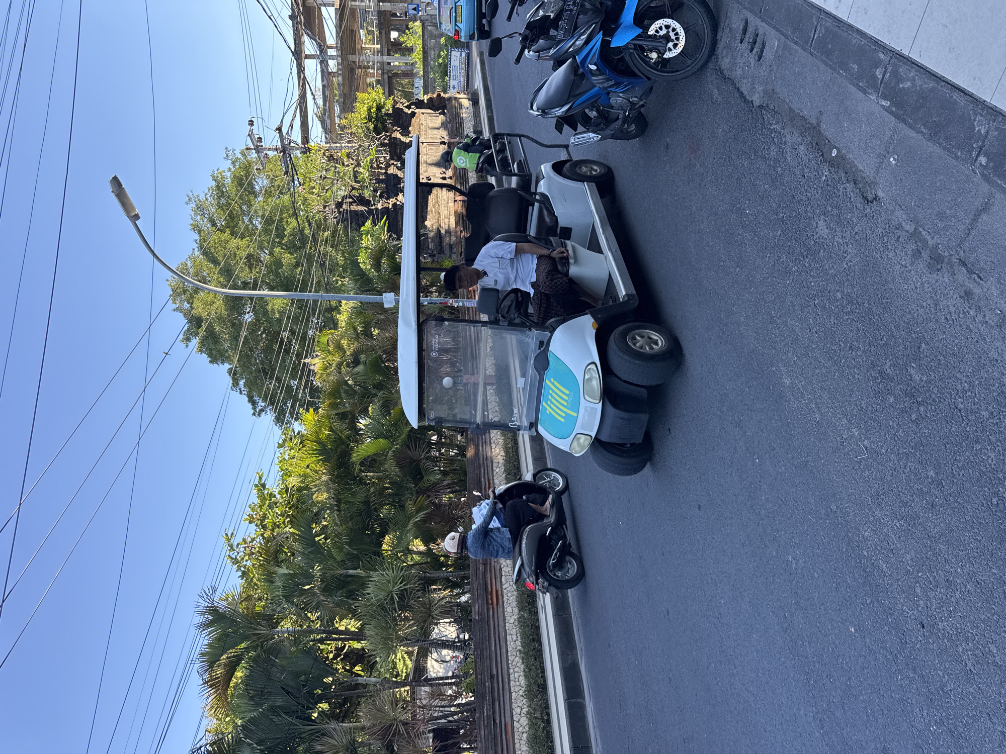 The image shows a street scene in Kuta, Indonesia. A white golf cart-like vehicle with the word "blluet" on the front is driving on the road. It appears to be passing a parked black scooter. The driver of the cart is wearing a white shirt and patterned trousers. A person on the scooter is wearing a denim jacket and a white helmet. A blue motorcycle is parked nearby as well. On the side of the road is a dense area of tropical vegetation and a weathered wooden fence, next to some train tracks. Overhead power lines and a street lamp are also visible. The sky is clear and blue.
