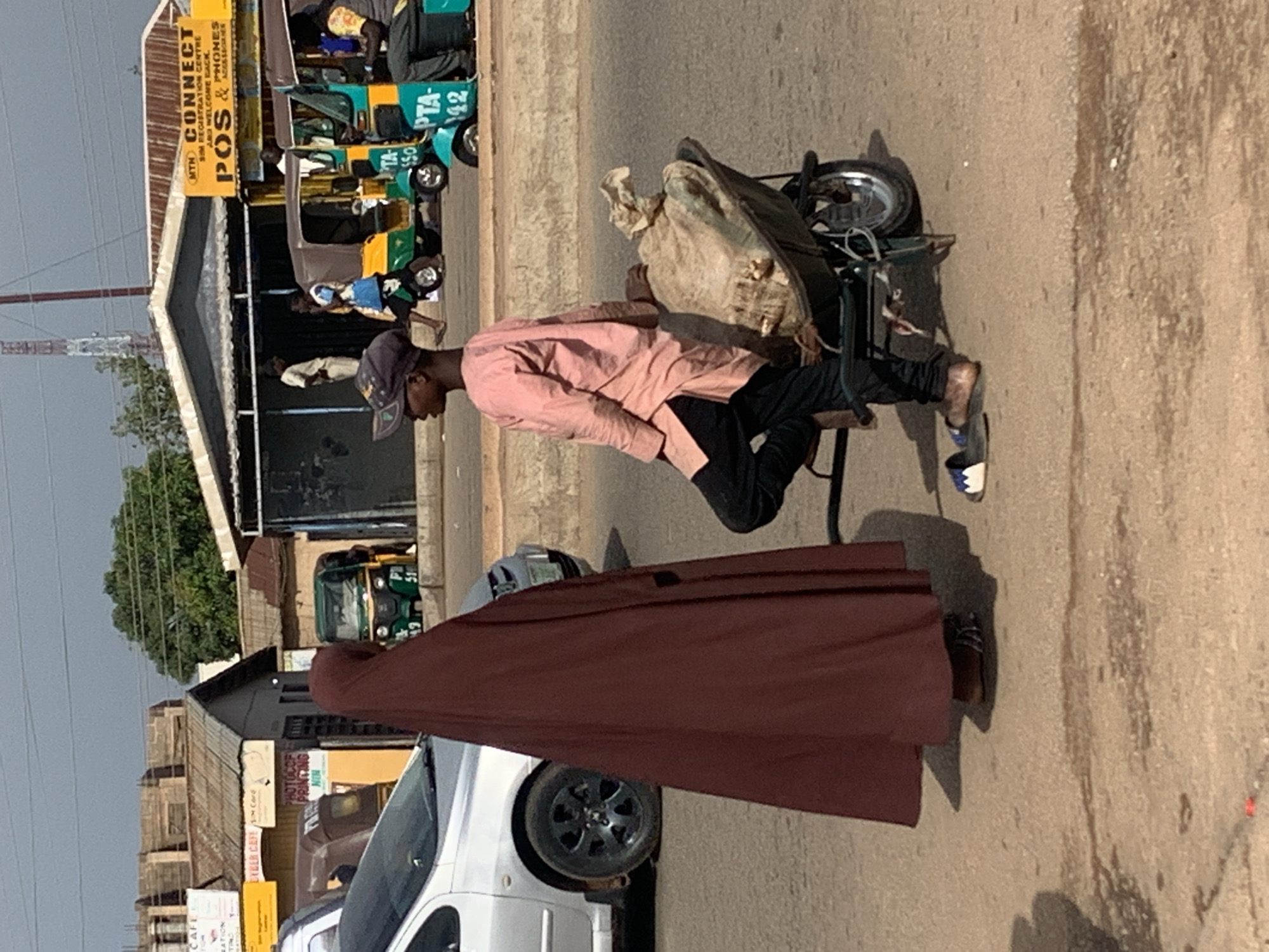 A young male wearing a pink shirt, dark pants, a baseball cap, and sandals stands next to a wheelbarrow filled with a large, light-colored sack. He is positioned on a paved road. To his left, a person wearing a dark brown hijab stands facing him. The background shows a street scene with buildings, vehicles including a silver car and several colorful auto-rickshaws, and trees under a cloudy sky. The location is identified as Gura Top, Nigeria.