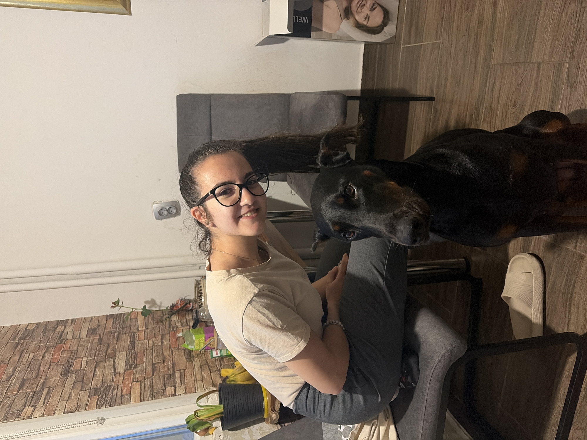 A young woman with dark hair, glasses, and a light-colored t-shirt sits in a gray chair. She smiles at the camera. A black Doberman stands beside her, partially obscuring her. The room has light-colored walls and wood-look tile flooring. In the background, there is a gray headboard and a box with a woman's face on it. The presence of a European-style electrical outlet suggests a European location. The presence of a light switch suggests the location is in Bulgaria, specifically Sofia. The items near the window, including the brick-patterned wall covering and the arrangement of objects on the shelf, provide further corroborating detail about the location.
