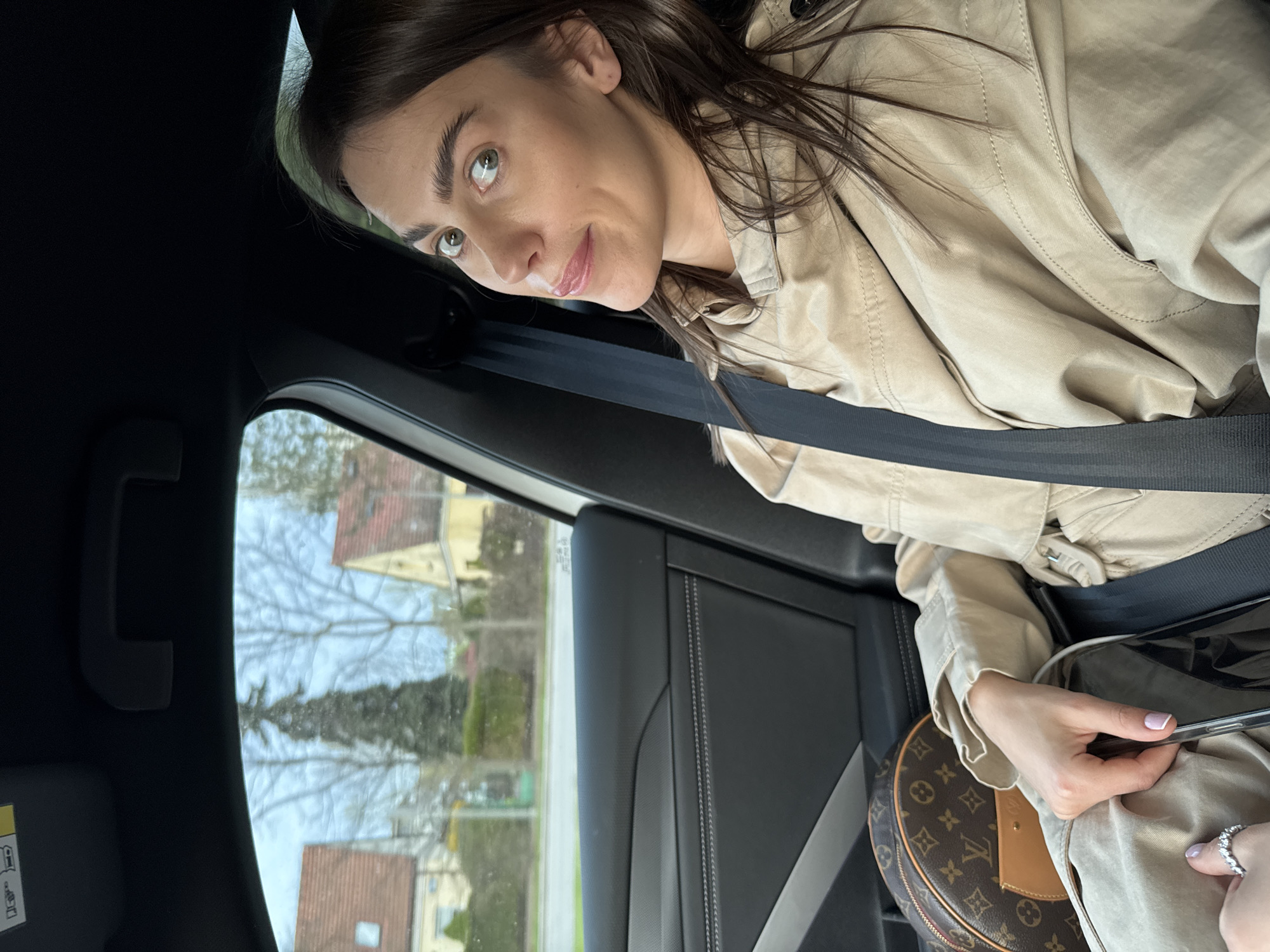 A woman is sitting inside a car, wearing a beige jacket and a seatbelt. She is holding a smartphone and has a small, round Louis Vuitton bag. The view through the car window shows a suburban street with houses and trees, possibly located in Warsaw, Poland. The scene appears to be during the daytime.