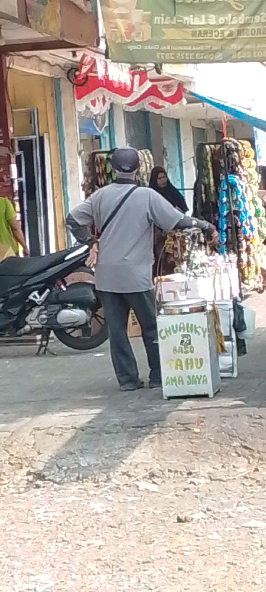 In Cilaku, Indonesia, on a paved street, a person wearing a grey t-shirt and a blue cap stands beside a food cart. The cart is white and displays a sign with green lettering that reads "CHUANKY BASO TAHU AMA JAYA". A black motorcycle is parked to the left of the cart. In the background, a woman wearing a black hijab is visible near a display of colorful items, possibly textiles. Other people and buildings are also present in the scene.