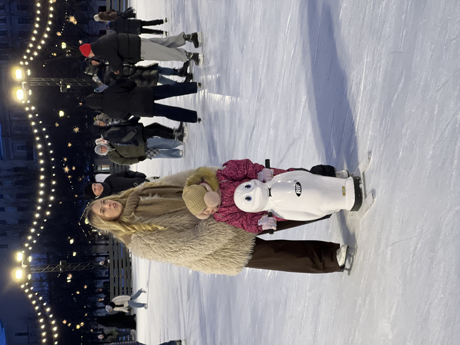 An outdoor ice skating rink in Saint Petersburg, Russia, is depicted at night, illuminated by overhead floodlights and decorative string lights. In the foreground, a young woman with long blonde hair, wearing a light beige faux fur coat, brown scarf, and dark brown trousers, skates on white ice skates. She stands slightly behind and to the left of a small child. The child, dressed in a bright pink padded snowsuit, a light brown knit beanie with a pom-pom, and pink earmuffs, holds onto a white penguin-shaped ice skating aid. The aid features blue eyes, a black bow tie graphic, and the letters 'RIF' on its chest, resting on two runners on the ice. Numerous other individuals, dressed in various winter attire and primarily on ice skates, populate the mid-ground and background of the rink, some actively skating and others standing. Dark, multi-story buildings are visible in the distant background against the night sky, behind a low barrier at the rink's edge.