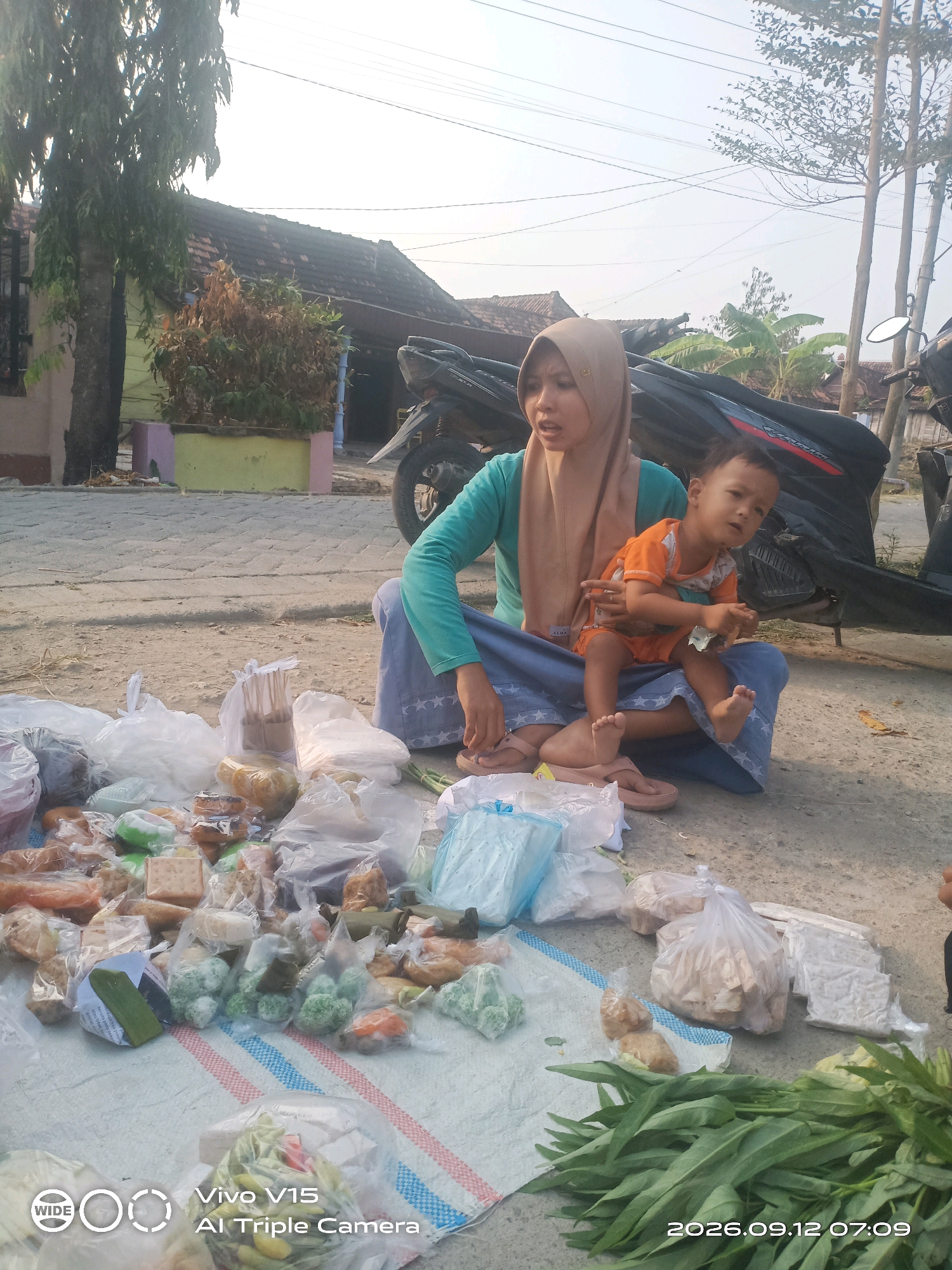A woman wearing a tan hijab and teal shirt is sitting on the ground in Plosorejo, Indonesia, holding a young child dressed in an orange shirt and shorts. They are surrounded by various food items packaged in plastic bags and on a white tarp, suggesting they are at a market stall. The ground is dirt and paved. In the background, there are houses, trees, and motorcycles. The lighting suggests it is daytime.