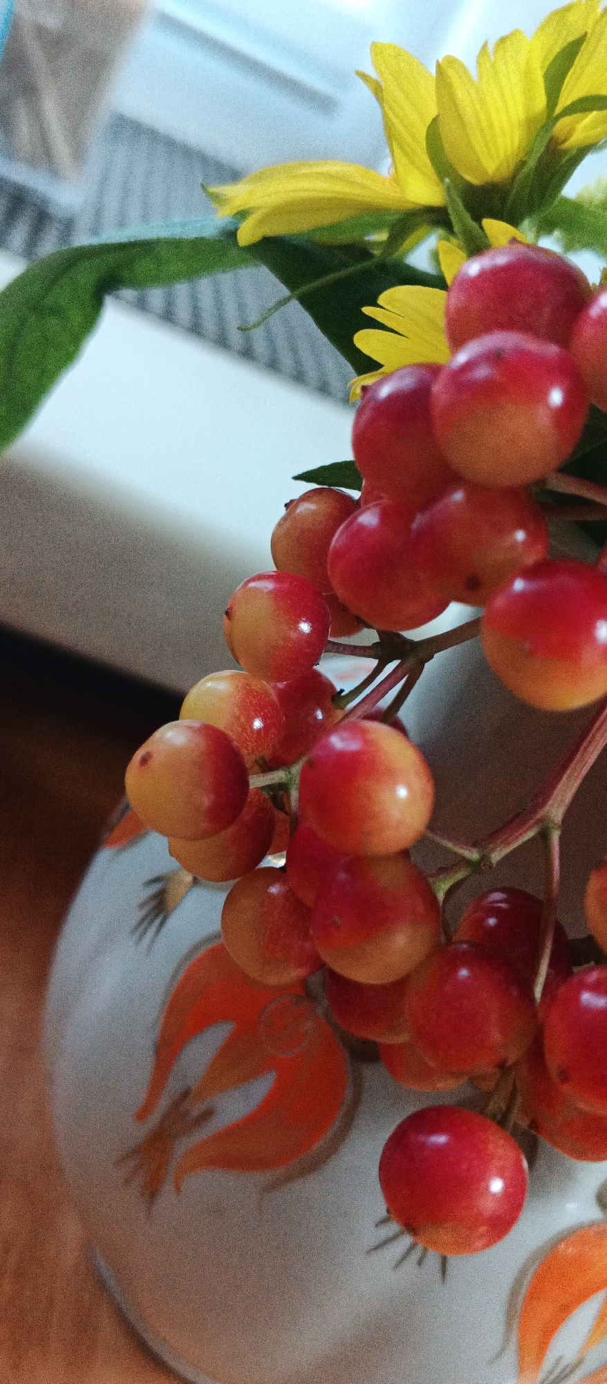 A close-up, vertical, indoor, natural light photograph shows a cluster of ripe red and yellow berries on a thin branch, positioned in the foreground to the right. The berries are spherical and glossy, some entirely red, while others display a gradient from yellow to red. In the upper right corner, a bright yellow flower with multiple petals is partially visible, also illuminated by natural light. To the left of the berries, a deep green leaf extends into the frame. The berries and flower are placed within a light-colored ceramic vase that has an orange floral pattern. The vase occupies the lower left quadrant of the image. The background consists of a blurred light-colored surface with horizontal lines, possibly a window blind or a table. The scene is domestic, and its location in Smila, Ukraine, is not discernible from the visual information presented in the image.