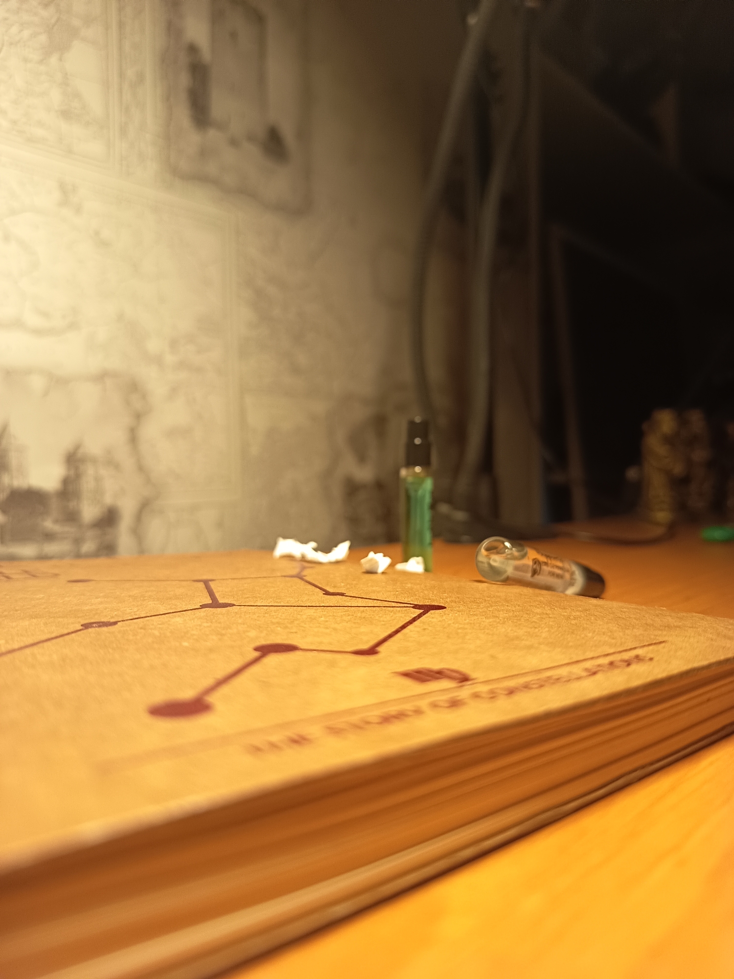 A low-angle, close-up shot focuses on a book with a diagram of constellations on its cover, resting on a wooden surface. To the right of the book, a small glass vial with a black cap lies on its side. Behind the book, a green vial with a black cap stands upright. Scattered on the book's cover are small, white, crumpled pieces of paper. The background is softly lit, showing a textured wall with faint patterns and blurred dark shapes, likely furniture or equipment. The image is taken indoors, likely in a domestic or office setting. There is no discernible information about the location in Izmail, Ukraine.