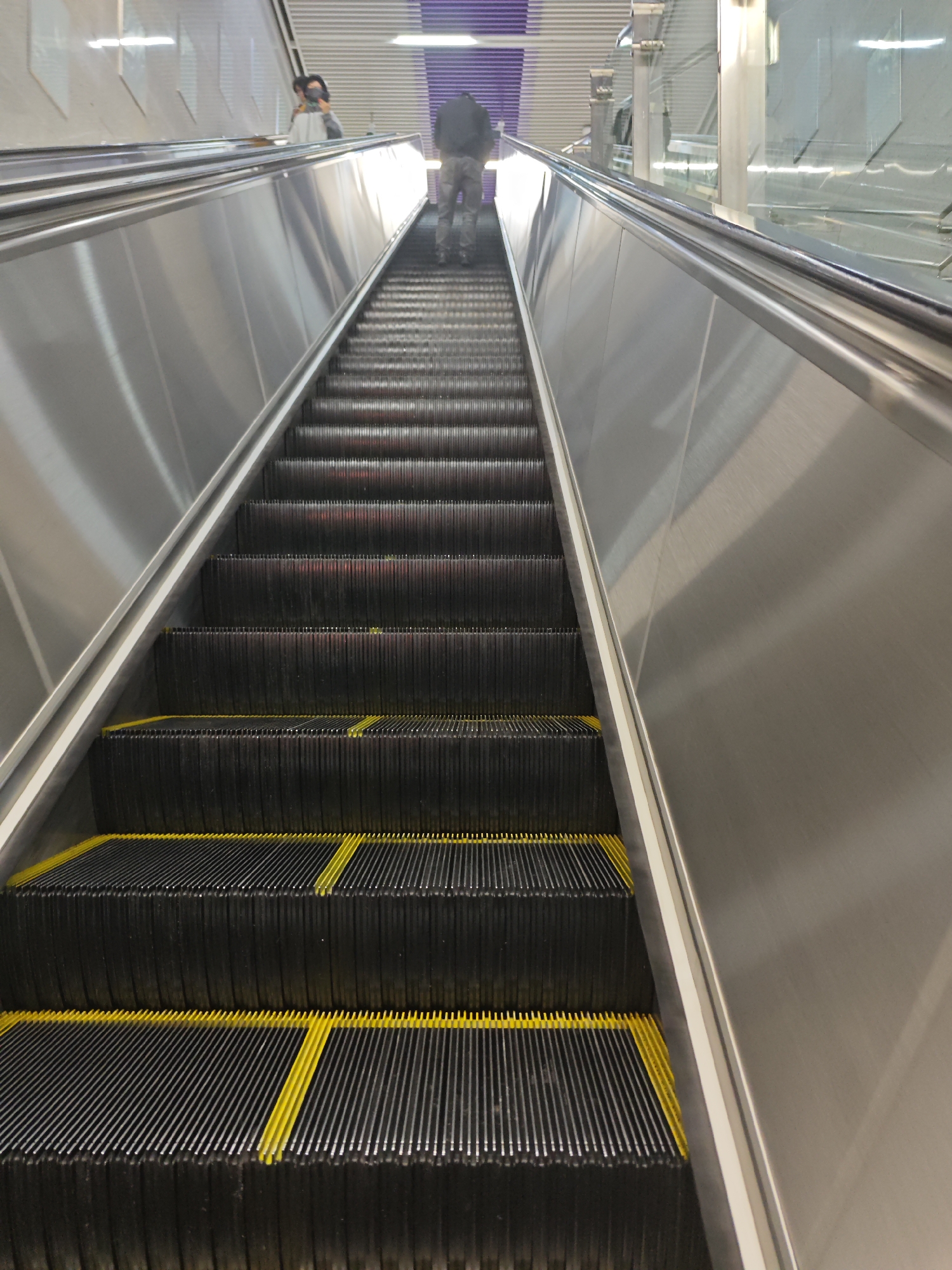The image shows an escalator in Liangjiang, China, moving upwards. The steps are black with yellow stripes. Two individuals are visible at the top of the escalator. The sides of the escalator are silver metal.