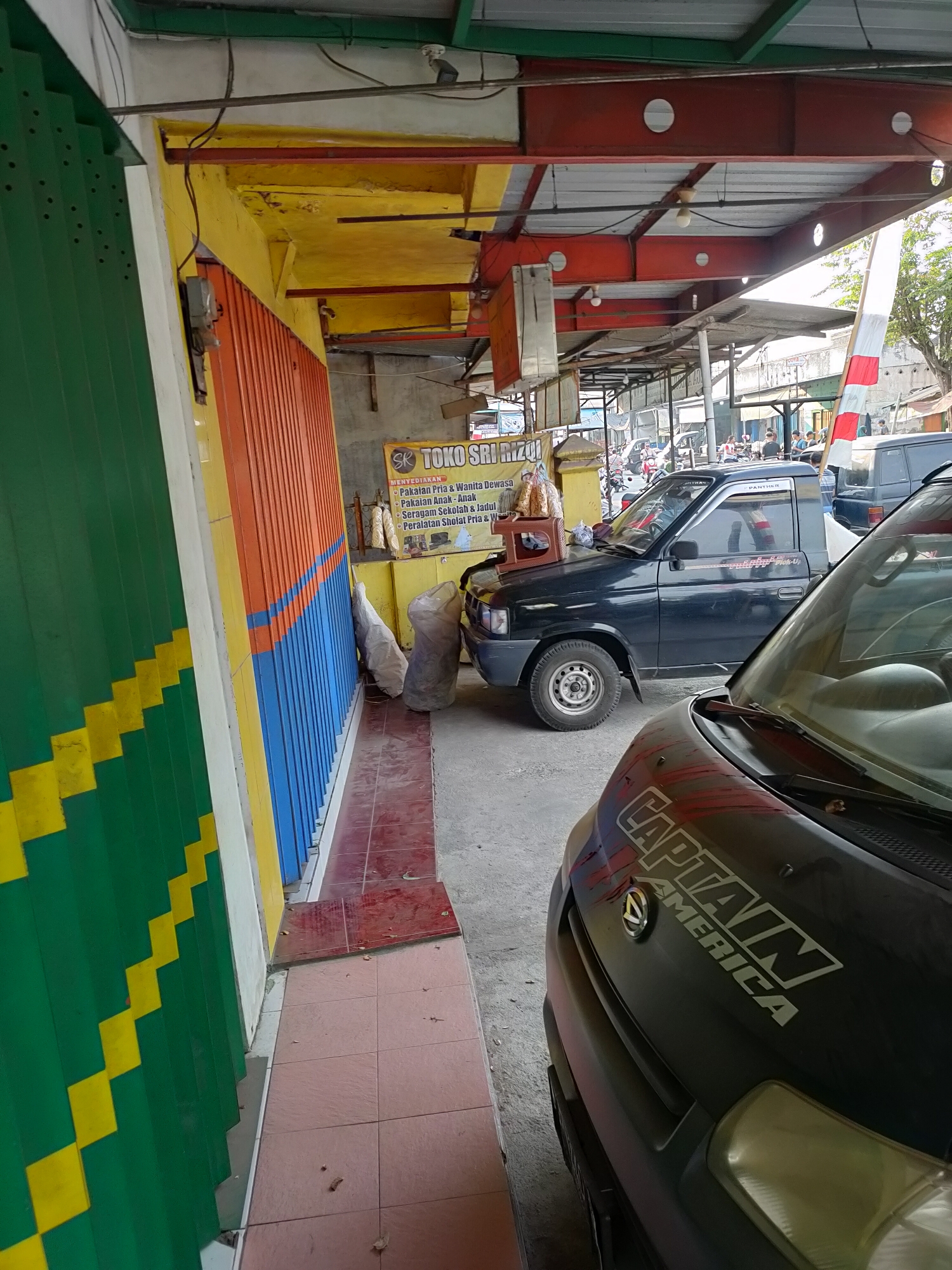The image shows a street scene in Ringinrejo, Indonesia, in front of a storefront. A green metal roll-up door with a yellow zigzag pattern is on the left. Adjacent to it is a wall painted in orange and blue vertical stripes. A sign for "TOKO SRI RIFAI" is visible, listing items such as men's and women's clothing, children's clothing, and school uniforms. Two large, white, tarp-covered bundles are leaning against the wall. A dark blue pickup truck with "PANTHER" written on the door and a white sticker on the windshield is parked in front of the store. In the foreground, the hood of a black vehicle with a white "CAPTAIN AMERICA" decal and a Daihatsu logo is prominent. The environment appears to be an open-air market or street stall area with a covered roof supported by red metal beams. Other vehicles and people are visible in the background.