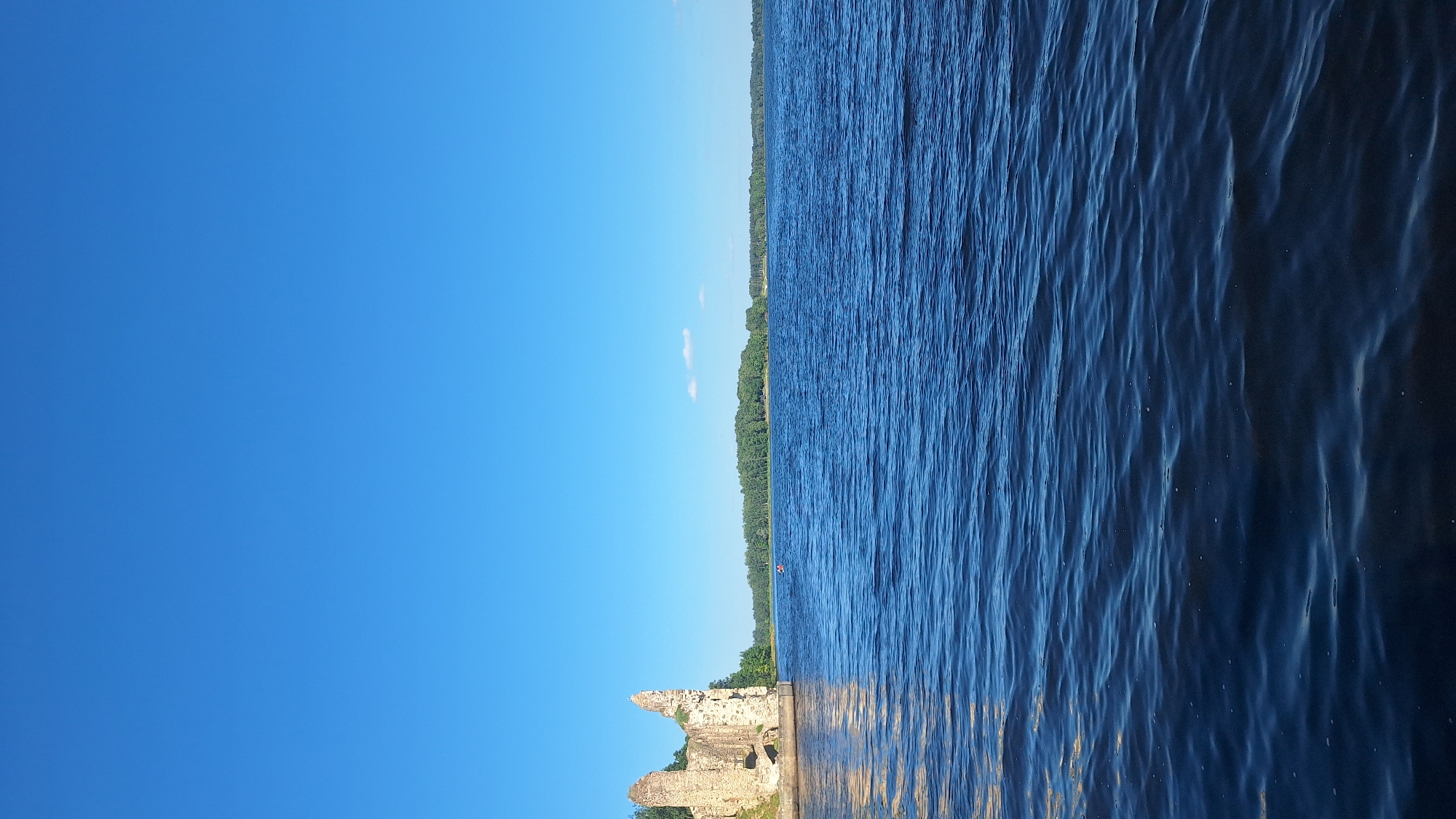 The image depicts a wide expanse of deep blue water, likely a river or lake, under a clear, bright blue sky. On the left side of the image, the ruins of an old stone structure, possibly a castle or fortress, stand prominently on a grassy embankment. The stone is weathered and crumbling, with sections of walls and towers still visible. Beyond the water, a dense, green forest lines the distant shore, creating a natural backdrop. The water is slightly rippled, suggesting a gentle breeze. Two small, red boats are visible in the middle distance on the water. The scene appears to be a historical site or natural landscape with remnants of past architecture. The location is likely Koknese, Latvia, known for its castle ruins situated on the Daugava River.