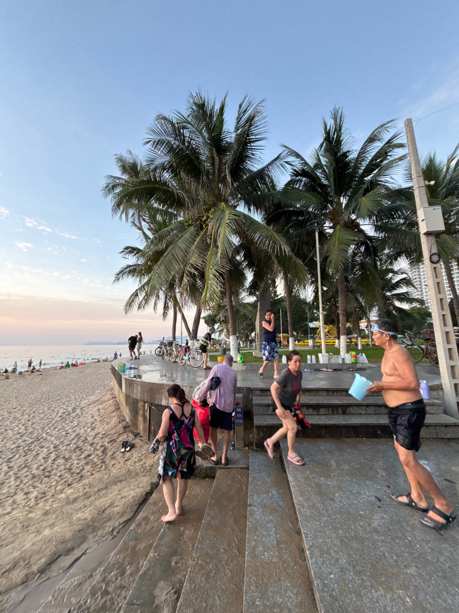 This image depicts a beach scene in Nha Trang, Vietnam. The foreground features a concrete promenade with several people walking on wet steps. A man in black shorts holds a light blue bucket, and a woman in a black and floral dress is walking down the steps. Further back, more people are visible on the promenade, some with bicycles. The background shows a sandy beach with numerous individuals enjoying the water and sun, with palm trees lining the edge of the promenade. The ocean stretches to the horizon under a clear, pale blue sky with some wispy clouds. In the distance, buildings and mountainous terrain can be seen. The overall atmosphere suggests a warm, late afternoon or early evening with recreational activities taking place.
