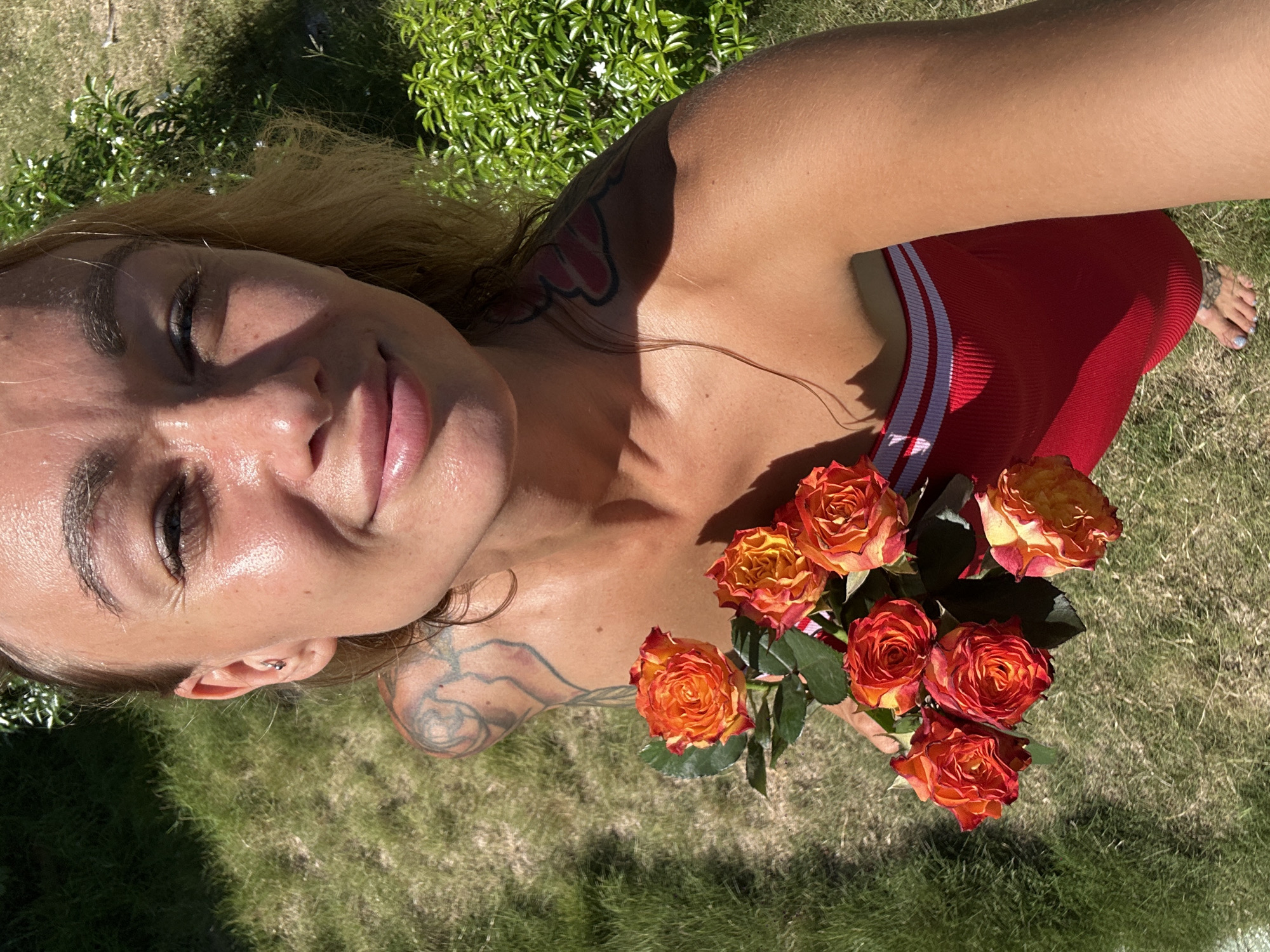 A young woman is shown from a high-angle perspective, holding a bouquet of orange and yellow roses. She is wearing a red strapless top with white stripes. Her hair is blonde and she has tattoos on her shoulders and arm. The background is out of focus and appears to be a grassy area with some green foliage. The lighting suggests it is daytime, with sunlight casting shadows. The image appears to be a selfie taken outdoors. The location is Mũi Né, Vietnam.