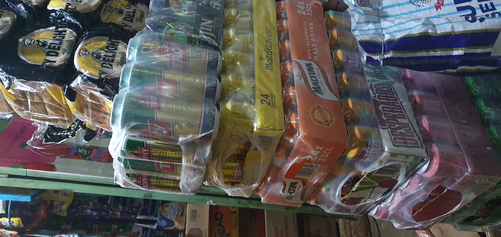 The image depicts a retail display of beverages and packaged food items in a market stall. In the foreground, stacks of drinks in cans are shrink-wrapped together. Brands visible include "Trophy," "Malta Guinness," and "Desperados." Some cans are gold and green, others are orange, and some are red. To the right, a large bag with blue and white stripes is partially visible, possibly containing snacks or other goods. Behind the beverages, there are packages of bread labeled "Y DELIGHT Bread." In the background, other market items are stacked, including boxes and bags of food products. The environment appears to be an indoor market stall, characterized by shelves and an overhead light source casting shadows. The scene suggests a commercial setting in Gura Top, Nigeria, where these goods are available for purchase.