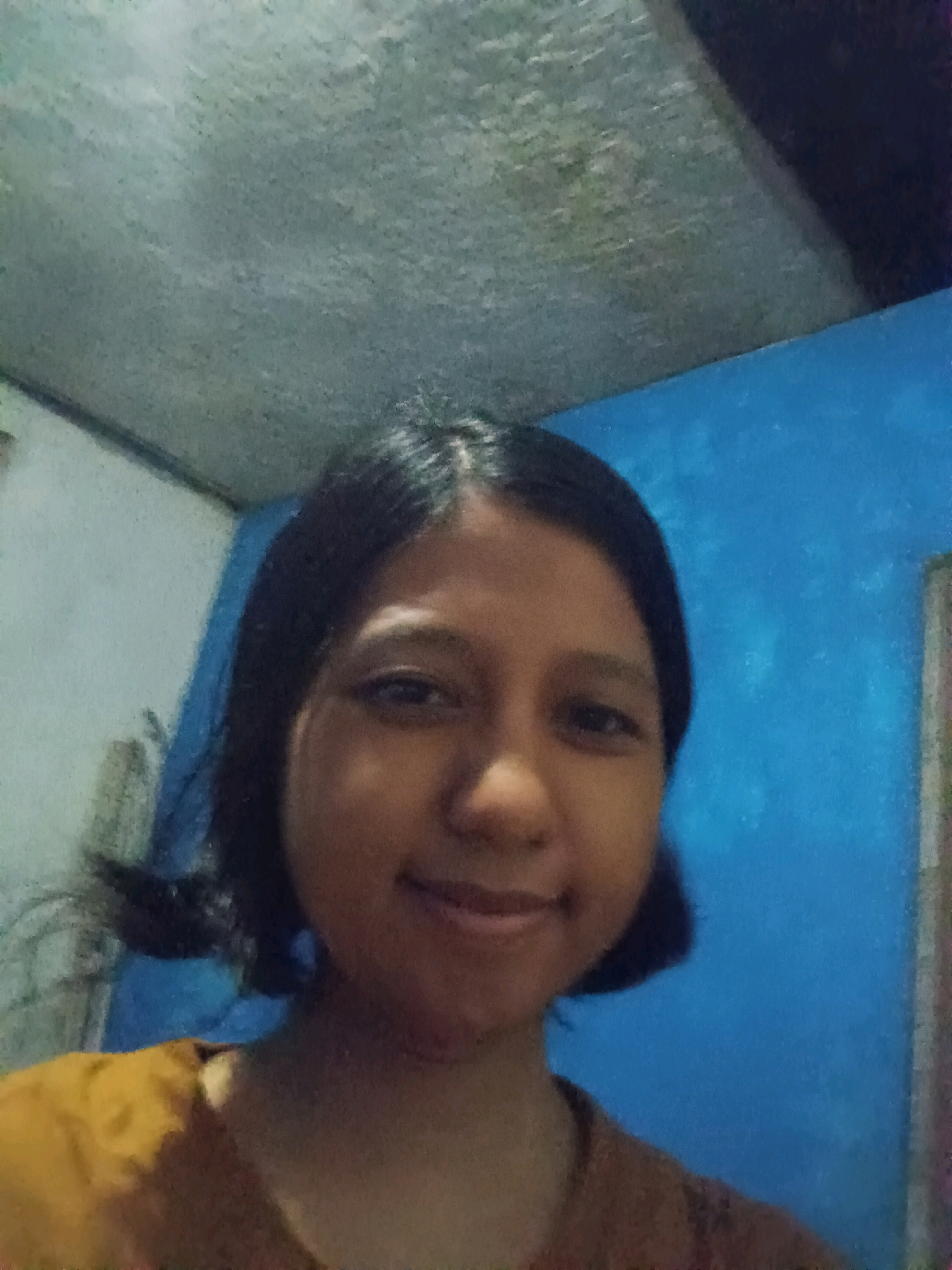 A young woman with dark hair pulled back into pigtails is captured in a close-up, frontal selfie. She is wearing a mustard-colored shirt. The background is a textured blue wall, and a portion of a gray ceiling is visible in the upper left corner. The lighting appears to be artificial and somewhat dim, casting a soft glow on her face. The setting is indoors, likely a room within a residential building in Canga'an, Indonesia, judging by the simple interior and the subject's likely ethnic background.