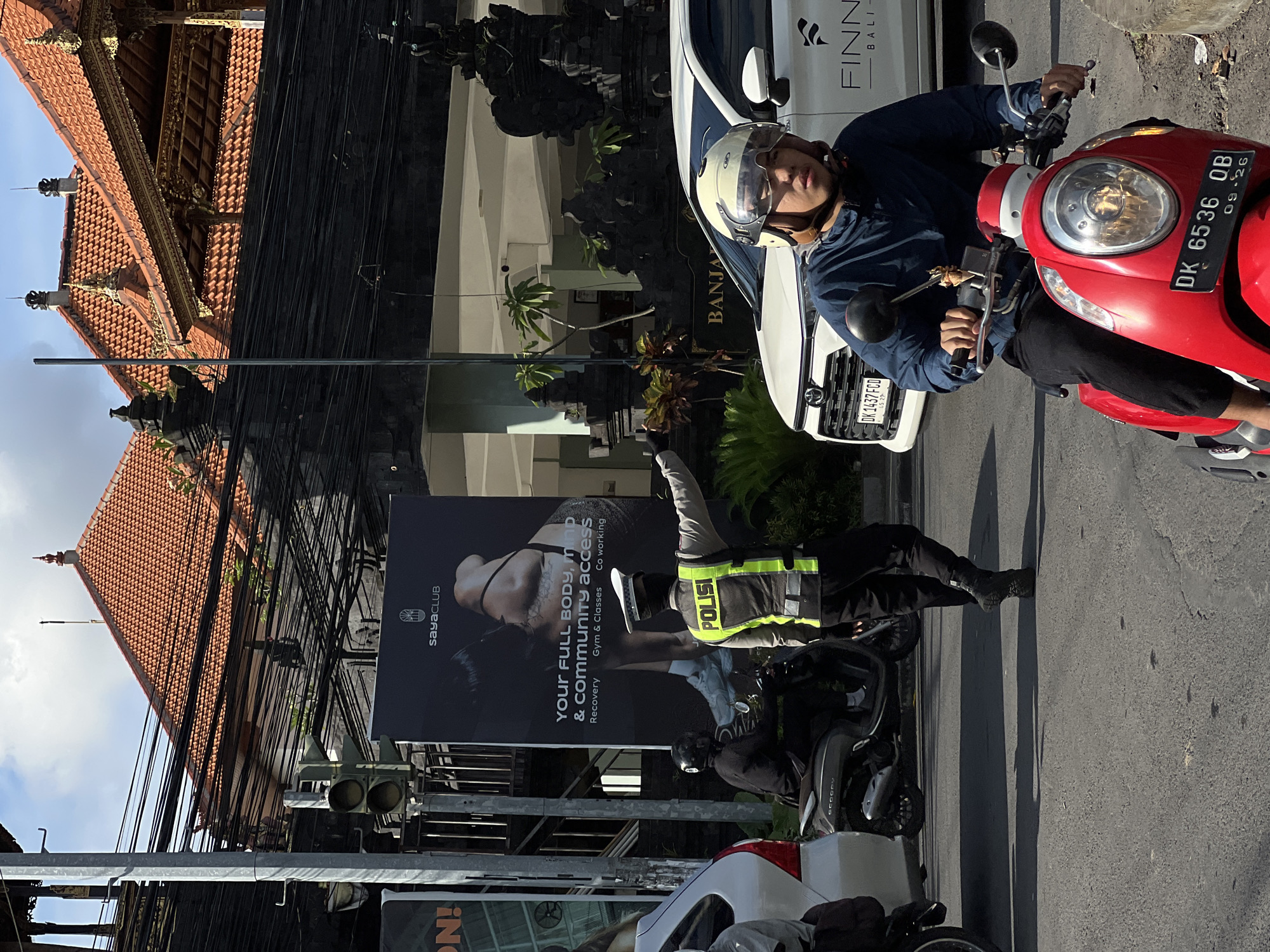 The image shows a street scene in North Kuta, Indonesia. A man wearing a helmet is on a red scooter with license plate DK 6536 OB. Next to him is a white car with the text "FINN BALI" and license plate DK 1437 FCD. On the left, there is a traffic signal, with numerous overhead power cables. A billboard advertises "sayaCLUB" with the text "Your FULL BODY, MIND & commUNITY Access," including services like Recovery, Gym & Classes, and Co-working. A police officer wearing a vest is standing next to a dark scooter. The roof of a Balinese-style building is visible in the background.