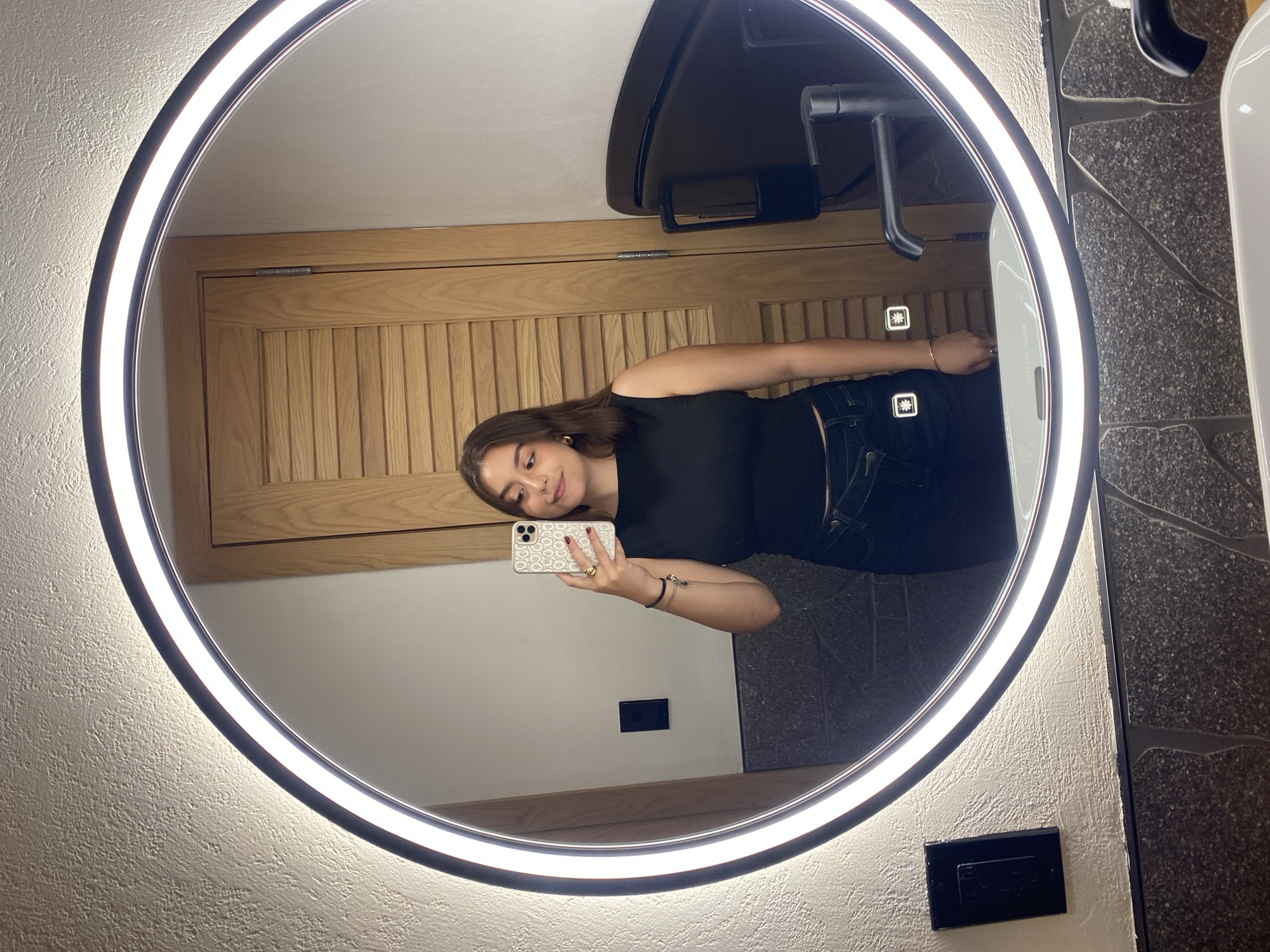 In Washington, United States of America, a young woman with brown hair is taking a selfie in a brightly lit bathroom. She is wearing a black sleeveless top and dark pants. She is holding a white and gold-patterned phone in her right hand. Her left arm is bent at the elbow, and she is wearing a bracelet on her wrist. In the mirror's reflection, a wooden door with horizontal slats is visible behind her. To her right, a black faucet and sink are reflected. The mirror is circular and has a bright LED light ring around its edge. The bathroom walls have a textured, light-colored finish, and the countertop appears to be made of dark, speckled stone.