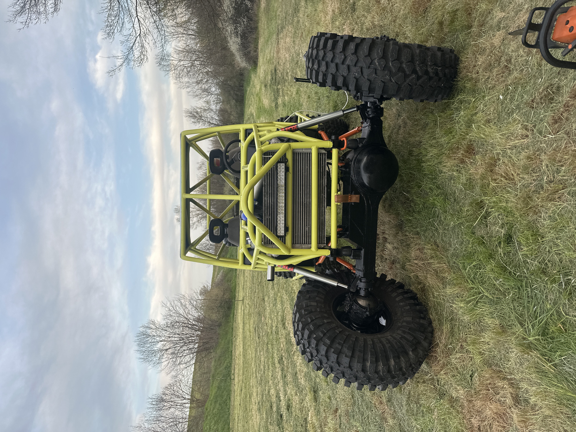 A yellow off-road buggy with large tires is positioned on a grassy surface. The buggy has a custom roll cage and visible radiator at the front. The background features trees, a blue sky, and some clouds. An orange chainsaw is partially visible in the upper right corner. The location is not specified as Choba, Bulgaria within the image.