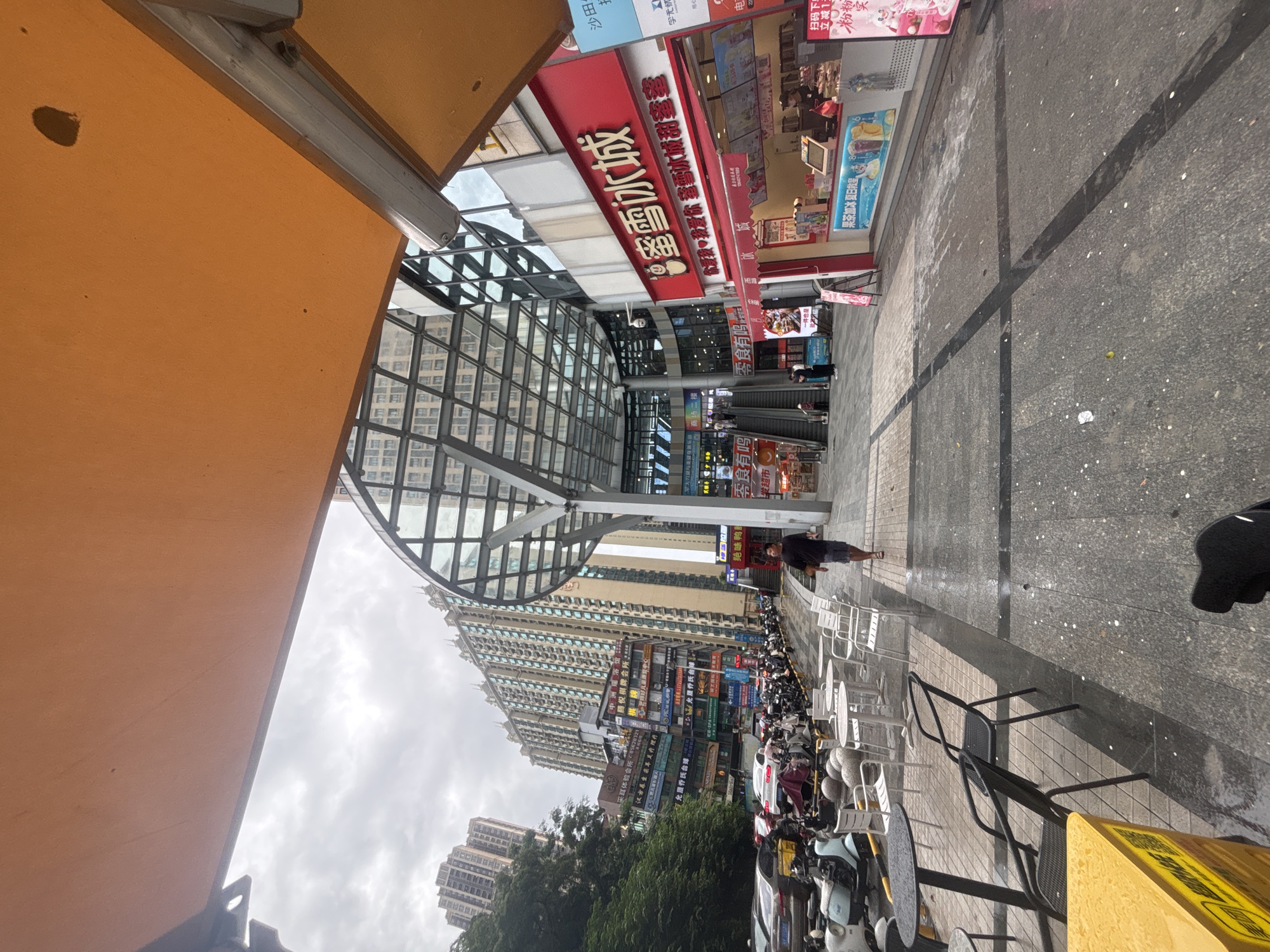 The image depicts a street scene in Nanhai District, China, likely in a commercial area. In the foreground, there are outdoor cafe tables and chairs. A person in a dark t-shirt and shorts is walking away from the camera on the right side of the pavement. Further down the street, an escalator leads into a modern building with a glass roof structure. To the right of the escalator is a red storefront with Chinese characters on its signage, appearing to be a food or beverage establishment. In the background, tall apartment buildings are visible, some with a reflective, glass-like facade. A row of parked motorcycles and scooters lines the left side of the street, along with parked cars. The ground surface is paved with a textured material, and there are visible wet patches and scattered debris. The sky is overcast.