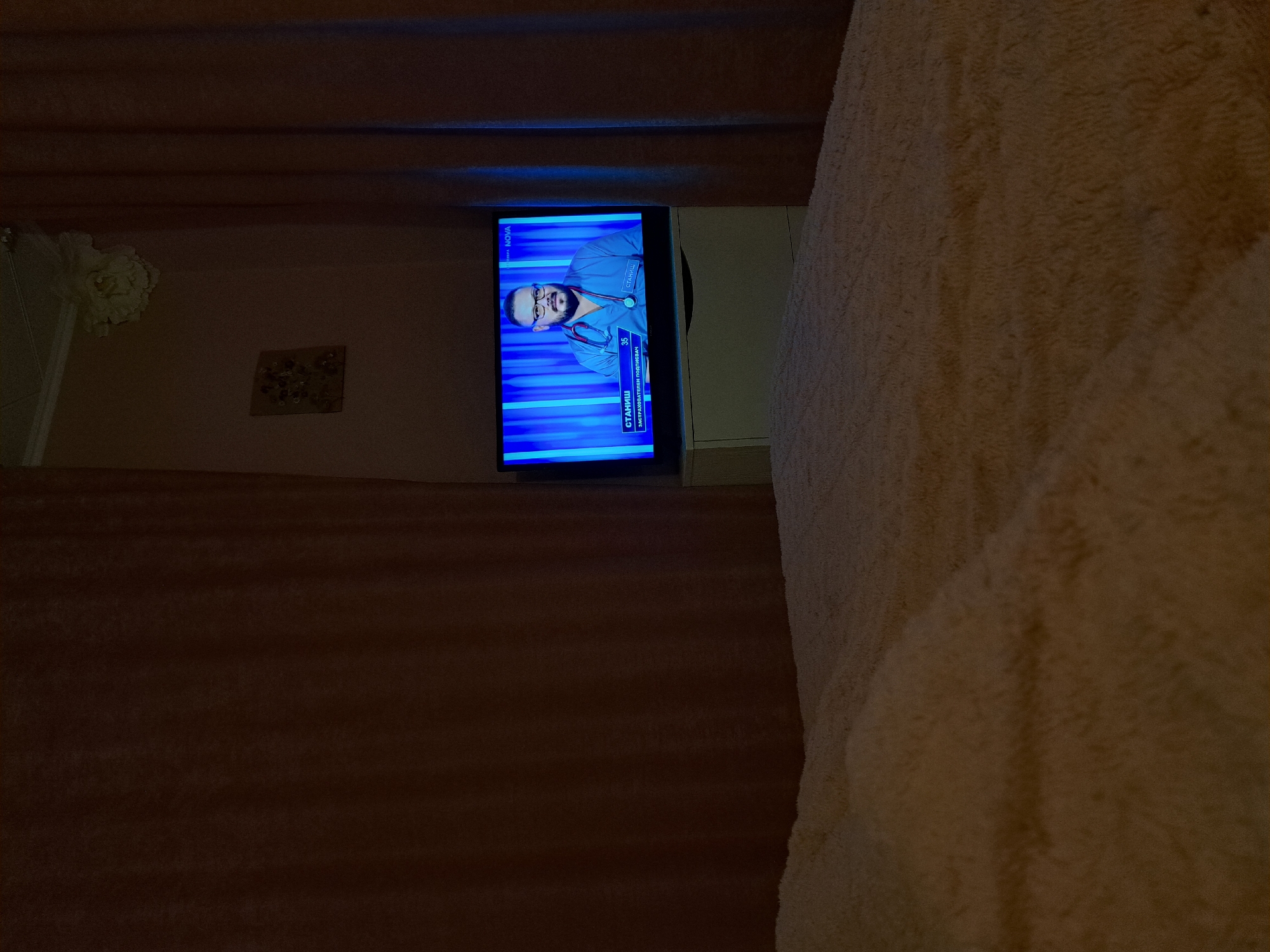 The image shows an interior setting, likely a room in a house or apartment. A television is mounted on the wall and displaying a man on the screen, presumably a doctor or healthcare professional. The TV appears to be tuned to a Bulgarian channel, as suggested by the text on the screen ("СТАНИШ"). The room is decorated with a light pinkish wall. A part of a sofa or a bed, covered with a light peach or tan-colored textured fabric is visible on the right side of the image. Additionally, there is an object resembling an ornament on the wall. The location, Chepintsi, Bulgaria, is not directly verifiable from the image.