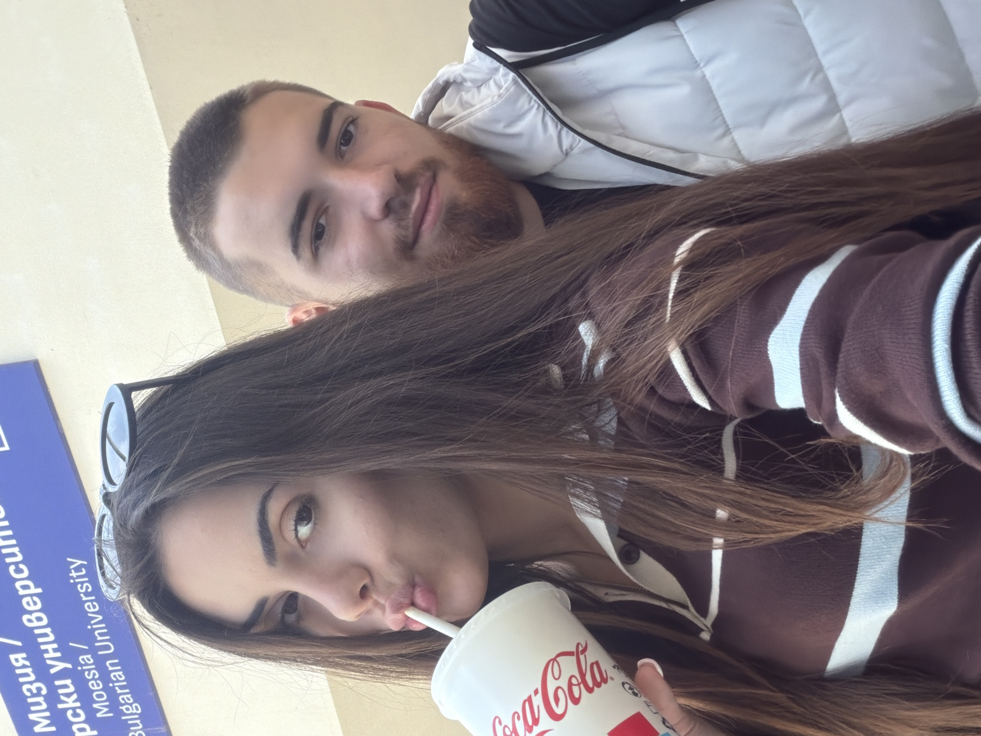 A young man and woman are posing for a selfie. The woman is in the foreground, drinking from a Coca-Cola cup with a straw. She has long brown hair and is wearing sunglasses on her head. She is wearing a brown sweater with white stripes. The man is behind her, wearing a white puffer vest and has short brown hair and a beard. A sign in Bulgarian and English identifies "Bulgarian University" which places this image near Sofia, Bulgaria.