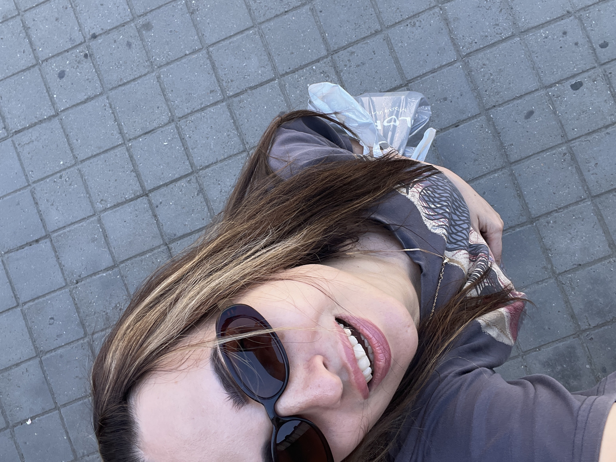 A high-angle, eye-level outdoor medium shot shows a woman with medium-length brown hair with blonde highlights, wearing dark sunglasses and a grey t-shirt with a graphic print. She is smiling with her mouth open and her teeth visible. She is holding a transparent plastic bag in her right hand. The ground beneath her is paved with grey square tiles.