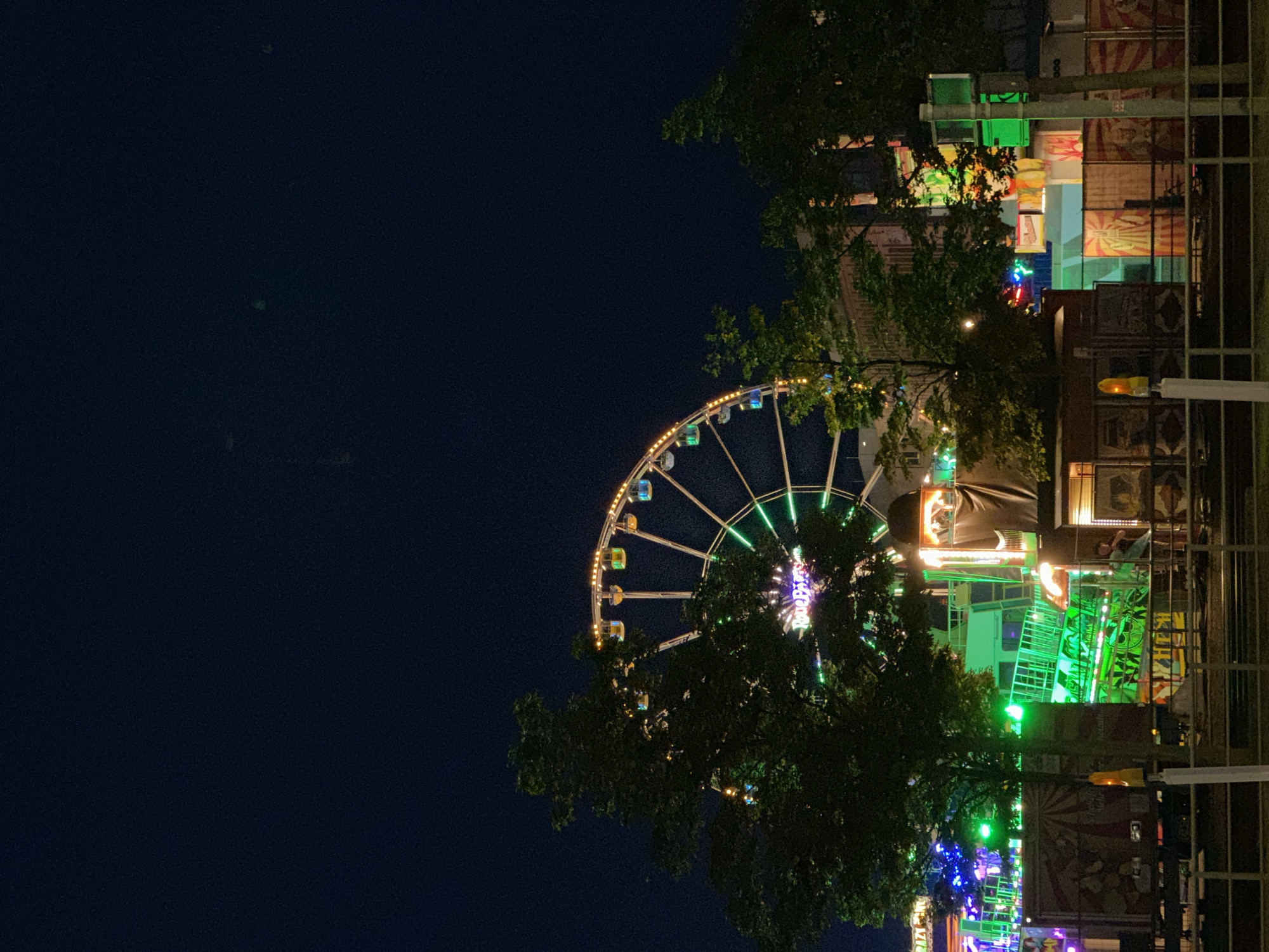 At night in the city of Kiel, Germany, a brightly lit Ferris wheel dominates the background of a temporary carnival or fairground. The Ferris wheel is adorned with numerous yellow and multicolored lights, with individual cabins also illuminated in various colors. In front of the Ferris wheel, trees with dense foliage partially obscure the view, their leaves catching some of the ambient light.
The foreground of the scene consists of carnival attractions and stalls. Structures with vibrant green and blue lights are visible, suggesting rides or game booths. Several transportable carnival units, some with colorful signage and graphics, are lined up. One unit features a large banner with red and yellow radial patterns. A person is visible standing near one of these structures, appearing to be an employee or attendee. Another person is seen further back within the illuminated green area. The overall environment suggests a lively event taking place after dark. A metal railing runs along the bottom of the frame, indicating a pedestrian area or boundary. The sky above is dark and clear.