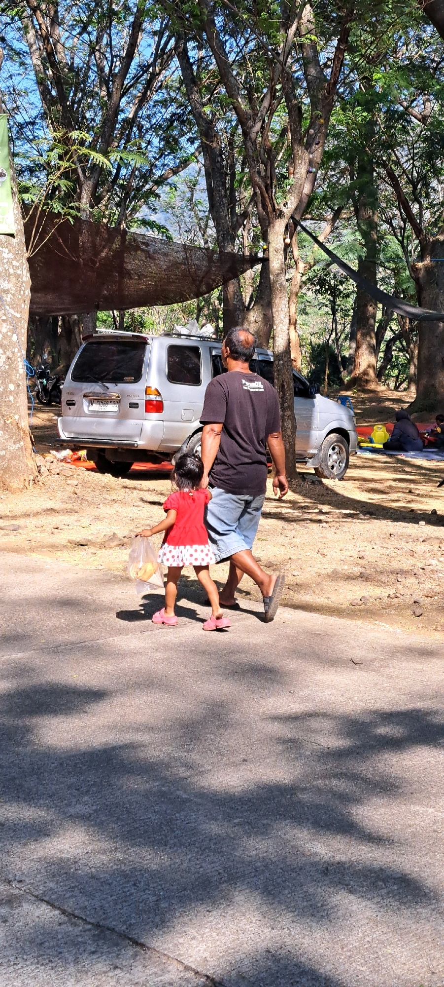 In Sumber Suko, Indonesia, a man and a young girl are walking on a paved road. The man is wearing a black t-shirt and denim shorts, while the girl is in a red and white polka dot dress and pink sandals. The girl is carrying a plastic bag with some food items inside. They are walking away from the viewer, towards the left side of the frame. Behind them, a silver minivan is parked under a canopy of trees. The environment appears to be a park or recreational area, with dense foliage and sunlight filtering through the trees. Other people are visible in the background, sitting on blankets.