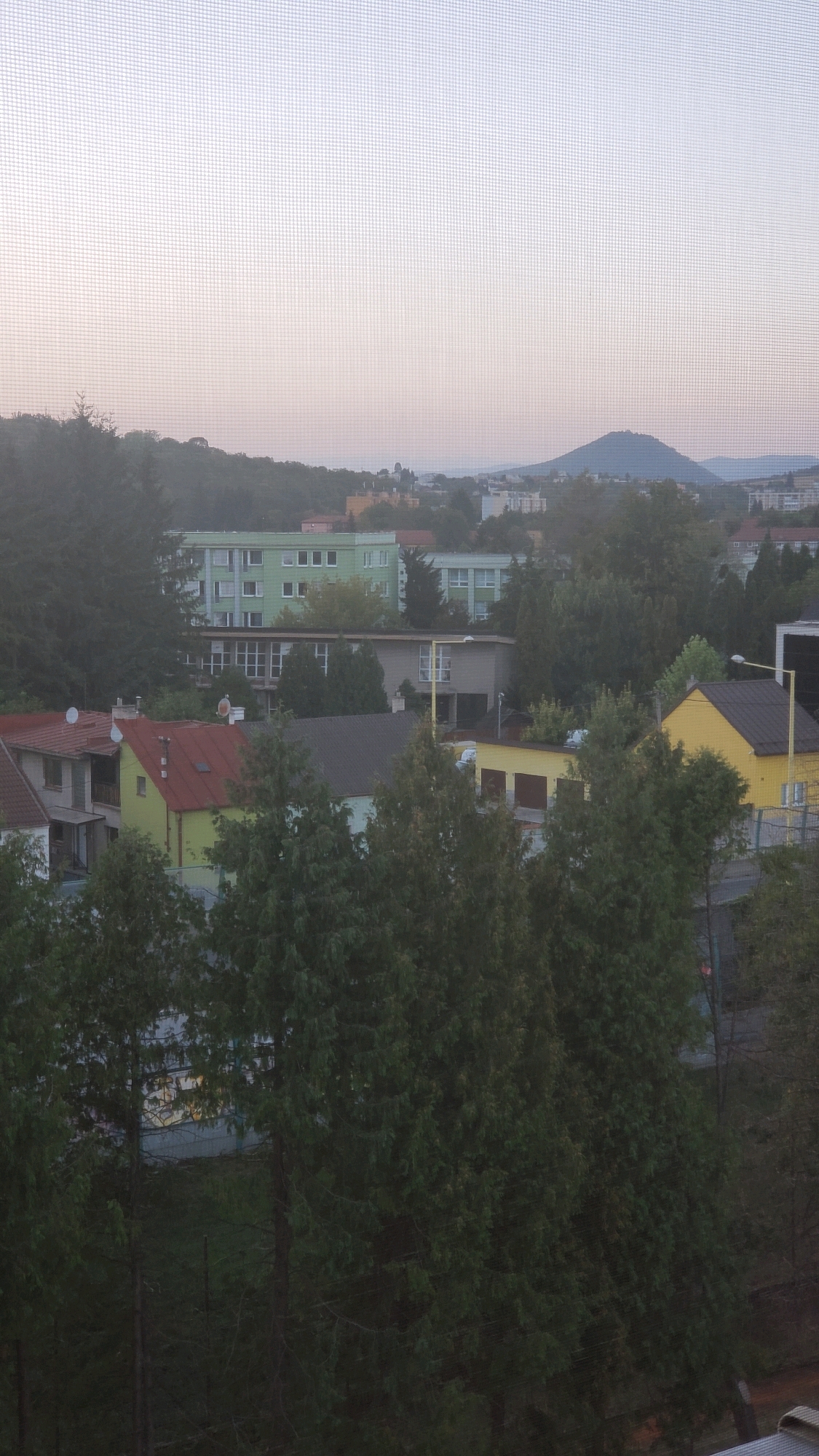 The image shows a view of the city of Prešov, Slovakia, from a window with a screen. The scene is a residential area with a variety of buildings and trees.
In the foreground, there are several houses. One house has a red roof, another has a bright yellow roof, and a third appears to be a light green color. These houses are surrounded by dark green coniferous trees.
Behind the houses, there are larger buildings, including a light green apartment block with many windows, and a beige building with a row of windows. Further in the background, more city buildings are visible, and a distinctive conical hill can be seen in the distance, characteristic of the landscape around Prešov. The sky above is a pale, hazy color, suggesting either early morning or late evening. The overall impression is a quiet, suburban or semi-urban setting. There are no people visible in the image. The focus is on the architecture and natural elements of the environment.