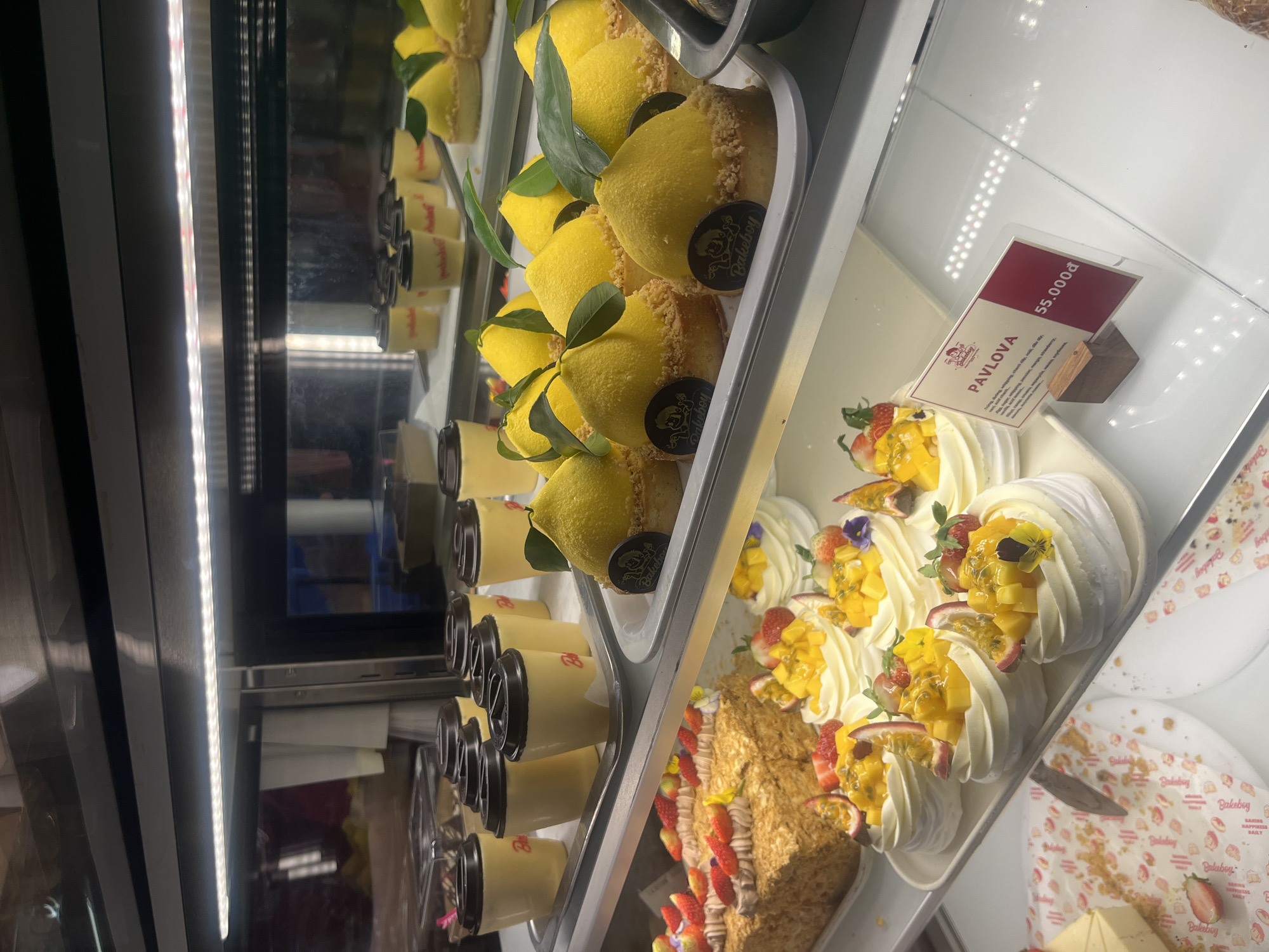 The image shows a display case filled with desserts at a Bakeboy location, likely in Vietnam due to the currency on the price tag. On the top shelf, there are lemon-shaped cakes and small cups of dessert, while the middle shelf holds meringue-based Pavlova desserts topped with various fruits like mango, strawberries, and passionfruit. The bottom shelf contains a layered cake with fruit and cream. A price tag for the Pavlova reads 55,000₫. The Bakeboy logo is visible on some of the desserts and packaging.