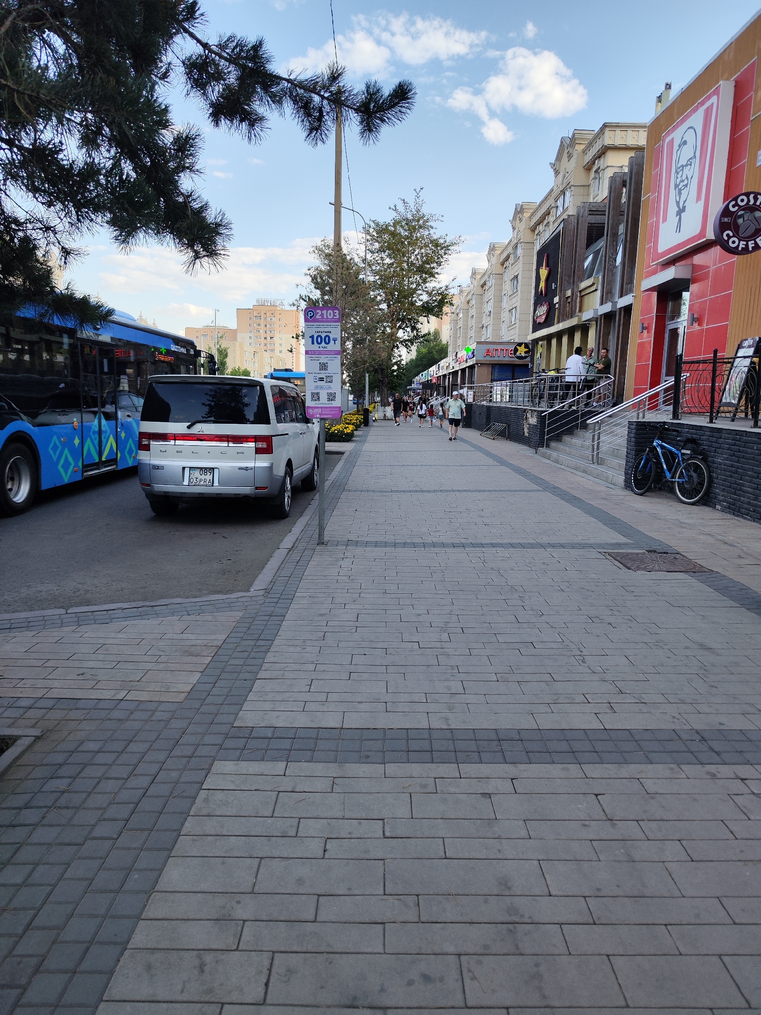 The image shows a street scene in Astana, Kazakhstan, on a sunny day. The foreground is dominated by a pedestrian walkway paved with grey and dark grey rectangular tiles arranged in a patterned fashion. To the left of the walkway, a grey asphalt road is visible, with a blue bus and a silver SUV parked on it. The SUV has a license plate with the characters "089 03 PRA". A lamppost with a parking meter attached is situated on the right side of the walkway.
Further down the street, several buildings line the right side, featuring a KFC restaurant with its distinctive red and yellow facade, and a Costa Coffee sign. There are people walking on the sidewalk and some are standing outside the establishments. Trees with green foliage are present in the background. The sky is blue with scattered white clouds.