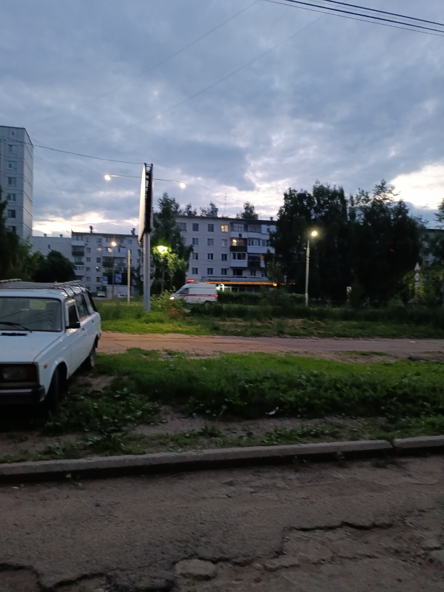 The image depicts a street scene in Chaikovsky, Russia, during what appears to be twilight. In the foreground, a white car with a roof rack is parked to the left. A paved road with visible cracks is in the immediate foreground, bordered by a concrete curb and a strip of grass. In the mid-ground, across a grassy area, is a road with a white van, possibly an ambulance or a utility vehicle, driving away from the viewer. Behind the road and grassy area are several multi-story apartment buildings, typical of Soviet-era architecture. Trees line the background, partially obscuring the buildings. Streetlights are illuminated, casting a warm glow. The sky is overcast with dark clouds. The overall impression is a quiet, residential urban environment at dusk.