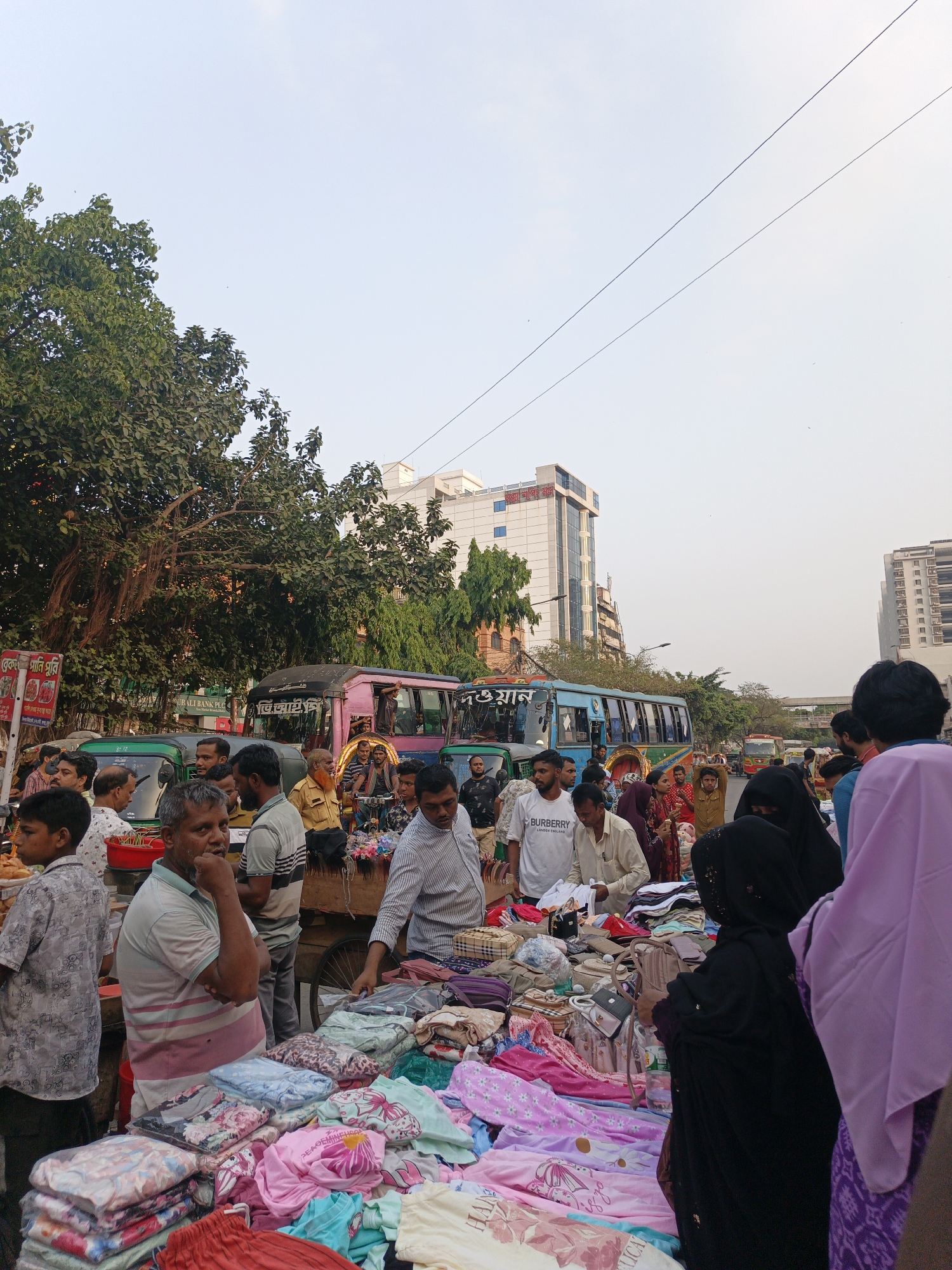 This image captures a vibrant and bustling street market scene in Dhaka, Bangladesh, likely during the late afternoon or early evening under a clear sky. The foreground is dominated by a street vendor's stall, laden with a colorful array of folded clothing items, including patterned garments that appear to be children's wear or casual textiles. Several people are gathered around the stall, either browsing the merchandise or engaged in conversation. A man in a striped polo shirt stands prominently near the goods, while another man wears a white t-shirt with "BURBERRY LONDON ENGLAND" printed on it.
The background reveals a busy urban environment with numerous people and vehicles. Public transport buses, including a distinctive pink bus with "ভিআইপি" (VIP) written on its front and a light blue bus, are visible, alongside auto-rickshaws. Large trees with aerial roots line the street on the left, and modern multi-story buildings rise in the distance, one clearly identifiable as "Rupali Bank PLC." Power lines crisscross the sky overhead.
Other visible text includes a red sign with Bengali script reading "ব্রেক পানি পুরি" (Break Pani Puri) and "দওয়ান," suggesting a food stall nearby. The overall scene depicts a lively commercial area filled with daily activities and interactions typical of a South Asian city market.