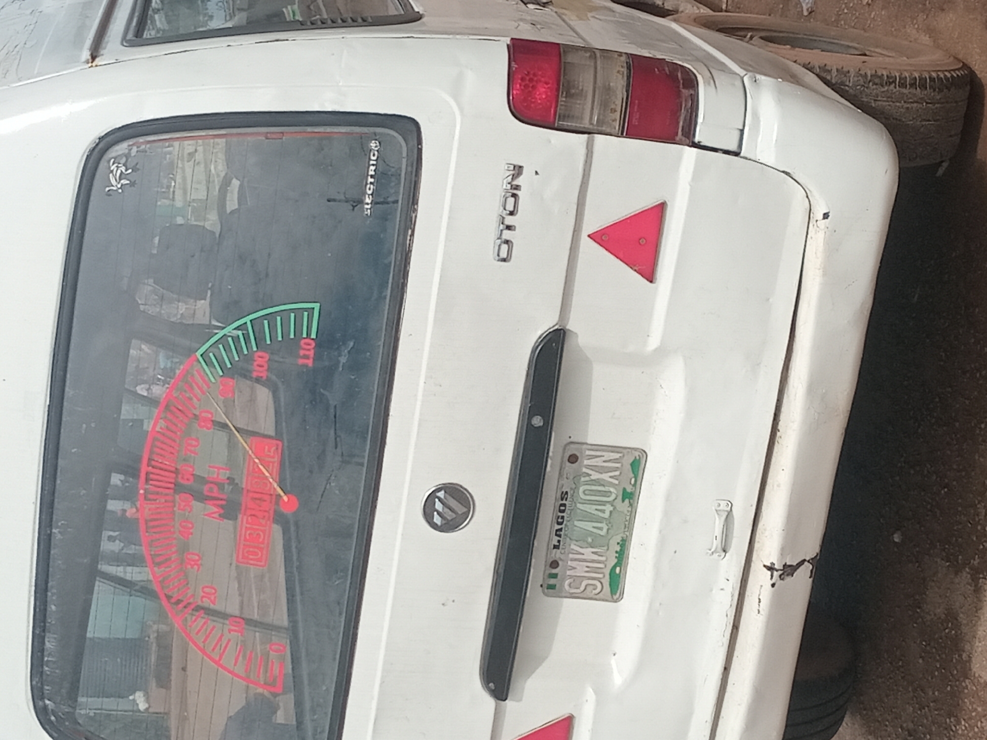 A white van, branded "OTON" on the rear, is parked on a dirt road. A circular metallic emblem with a wing-like design is centered above the license plate. The license plate, from Lagos, reads "SMK 440XN". A red triangular reflector is affixed to the rear of the van. A speedometer decal is visible on the rear window, showing speeds in MPH, with a needle pointing to approximately 45 MPH. The van has minor dents and scratches on its rear bumper and body. The surrounding environment is unpaved and appears to be an outdoor area in Bukuru, Nigeria.