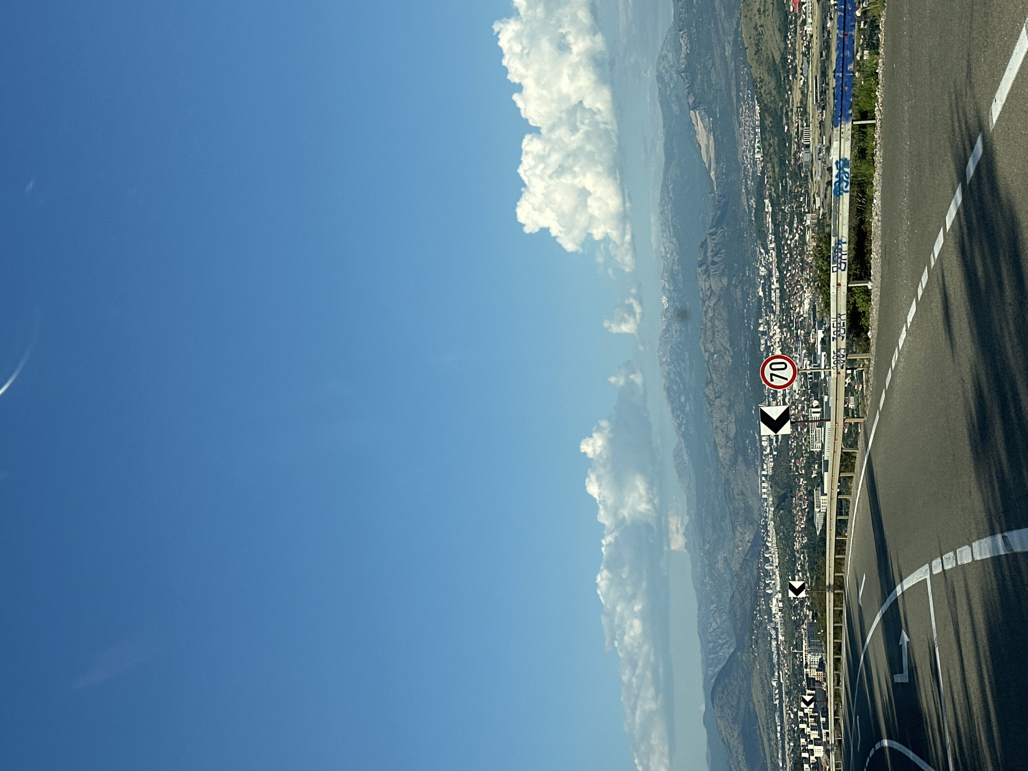 The image depicts a road with a 70 speed limit sign, a city view in the background, a mountain range in the distance, and a blue sky with white clouds. The location is Gornji Kokoti, Montenegro.