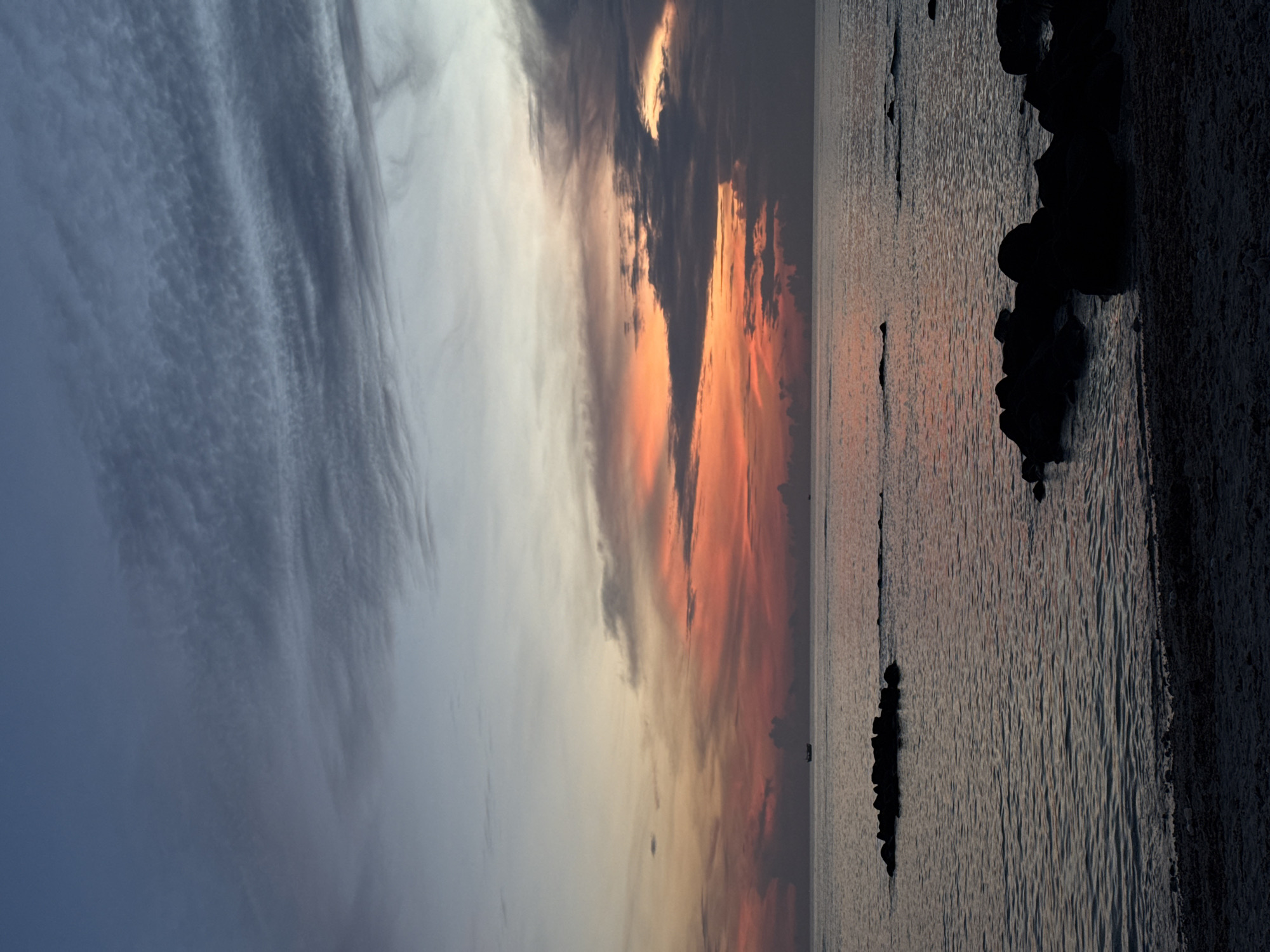 The image depicts a serene sunset over the ocean, likely taken from the shores of Lý Sơn Island, Vietnam. The sky is a gradient of blues and oranges, with wispy clouds creating a textured effect. The ocean is calm, with gentle ripples reflecting the colors of the sky. In the distance, a small boat is visible, adding a sense of scale to the vastness of the sea. Several dark, irregular rock formations emerge from the water, breaking the otherwise smooth surface. The foreground shows a dark, pebbly shoreline, with more rocks scattered along the water's edge. The overall atmosphere is tranquil and visually captivating, characteristic of a sunset by the sea.