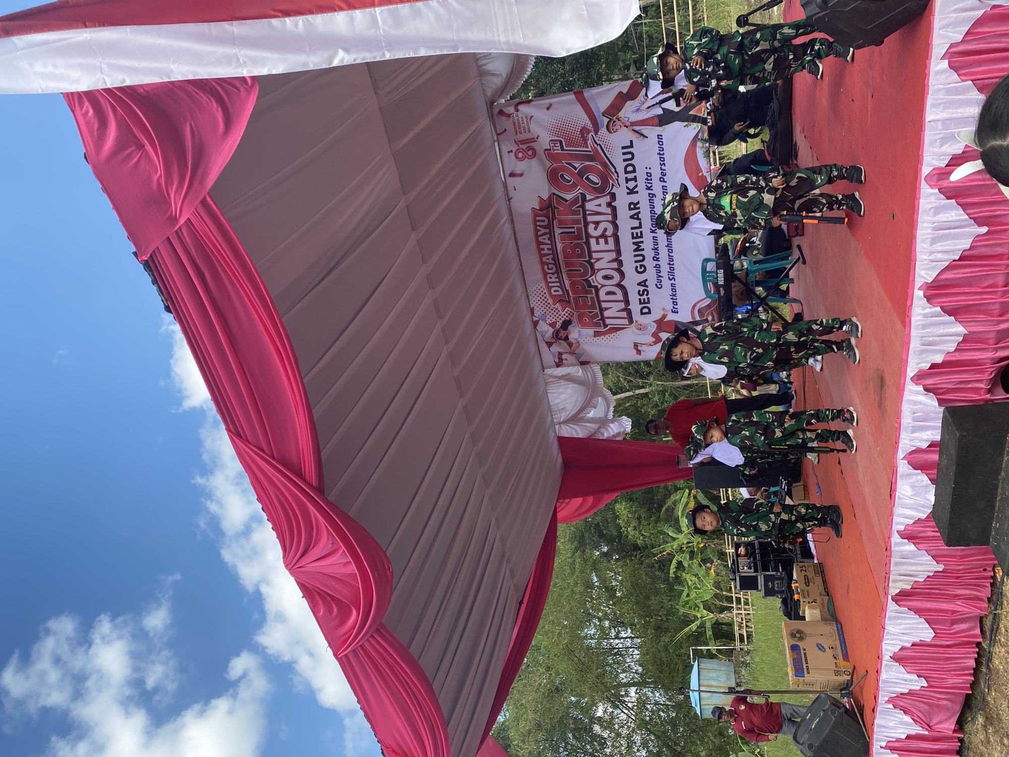 A group of children dressed in camouflage uniforms are standing on a red-carpeted stage under a pink and white tent. They are holding toy rifles and wearing camouflage hats. The backdrop behind them is a banner that says "DIRGAHAYU REPUBLIK INDONESIA 81" and "DESA GUMELAR KIDUL GUYUB RUKUN KAMPUNG KITA ERATKAN SILATURAHMI DAN PERSATUAN." To the left of the stage, there is a large black speaker, a fan, and several boxes. A man in a red shirt is standing to the left of the stage, looking towards the children. Another man in a red shirt is standing behind the tent on the left side of the image. The environment appears to be outdoors, with trees and greenery visible in the background. The sky is blue with scattered clouds. The scene is likely at a community event in Desa Gumelar Kidul, Tambak, Indonesia, celebrating Indonesian Independence Day.