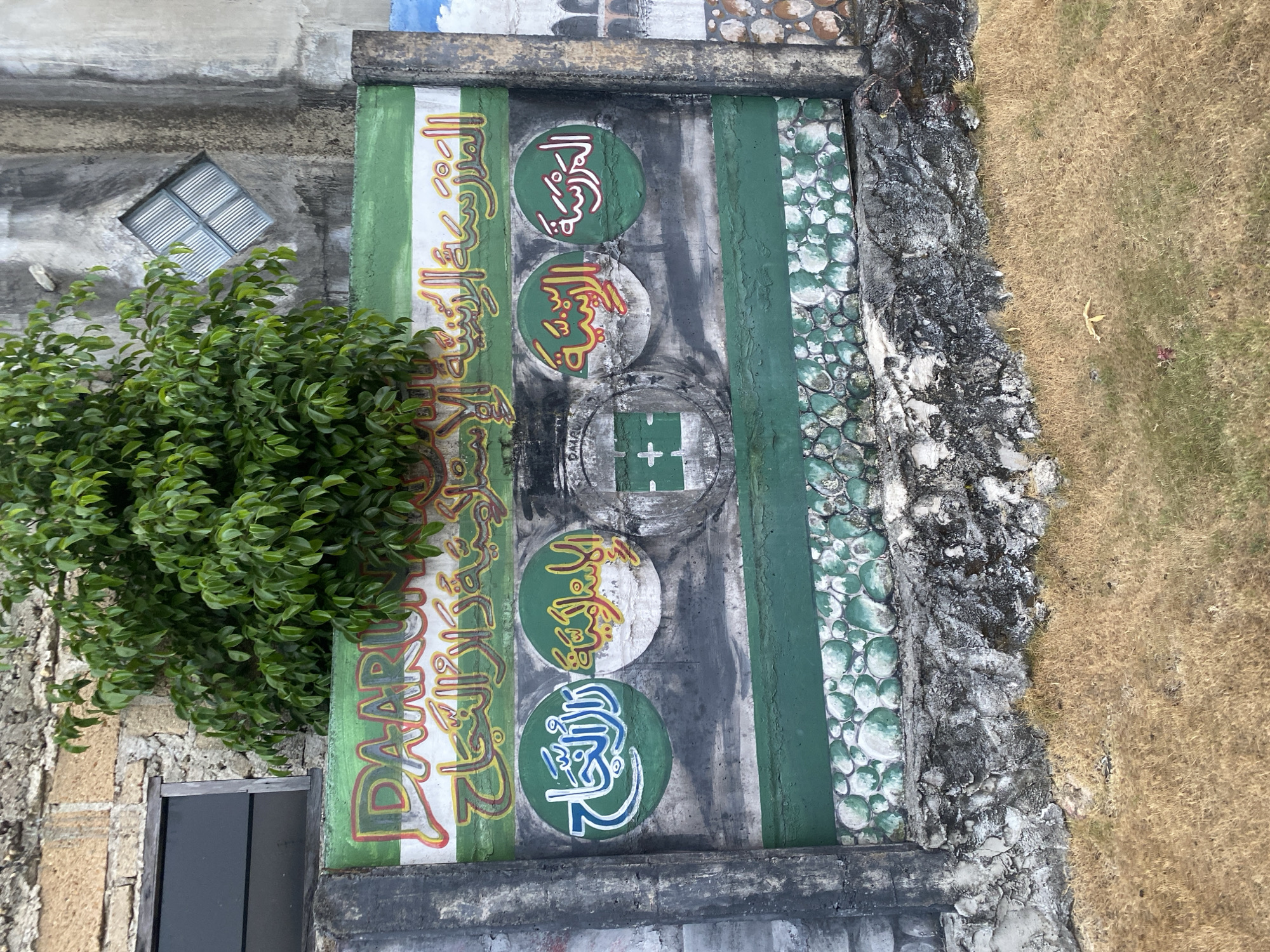 The image shows a colorful mural painted on a concrete wall in Banjarbaru, Indonesia. The mural features Arabic calligraphy, the name "DAARUL ULUUM" in red and white letters, and several circular emblems with Arabic script. A green and white striped pattern runs across the top of the mural, and a row of painted green and white stones is visible at the bottom. A large green bush grows from behind the wall, obscuring part of the mural. To the left of the mural, a dark-colored window is set into a stone wall. To the right, the concrete wall continues and shows signs of weathering. In the foreground, dry, golden-brown grass covers the ground. The overall scene suggests an outdoor environment, possibly the exterior of a school or religious building.