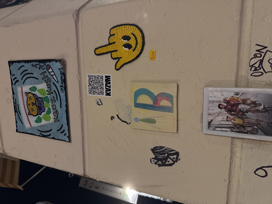 The image depicts a section of a light-colored building wall, likely a pillar or corner, adorned with multiple pieces of street art, stickers, and graffiti. The surface shows minor wear, including areas of chipped paint and surface texture. The scene is illuminated, suggesting nighttime or indoor ambient lighting conditions within the city of Paris, France.
From top to bottom, left to right:
* A rectangular sticker or small print features a stylized cartoon cat's head with "X" eyes and purple drool, enclosed within a clear plastic bag graphic. Green cannabis leaves surround the cat. Below this, the text "Kaoz Kush" is printed, accompanied by an illustration of a lit joint. The artist's handle "@kaozart" is visible at the bottom.
* Below this, a black and white QR code sticker is present, with the word "KVZVM" in white text on a black bar beneath it.
* To the immediate right of the QR code, a yellow, textured sticker or artwork depicts a human hand making a 'middle finger' gesture. A smiley face is visible on the palm. A small yellow rectangular sticker with red text, possibly "Le Bo'm", is affixed below this.
* Below the QR code, a small rectangular artwork features a minimalist green stick figure on the left, beside a large, stylized letter "B" rendered in blue and red tones.
* To the left and slightly below the "B" artwork, a small, dark, abstract heart-shaped sticker with a patterned design is visible.
* Near the bottom center, a vertical rectangular print or photograph depicts three individuals in an urban street. One person is riding a bicycle, another is seated on the handlebars, and a third person in plaid trousers, carrying a bag, walks alongside them. The background shows old European-style buildings.
* At the bottom left, a partial graffiti numeral "9" is visible.
* At the bottom right, fragmented graffiti spelling "ORSON" is partially visible.