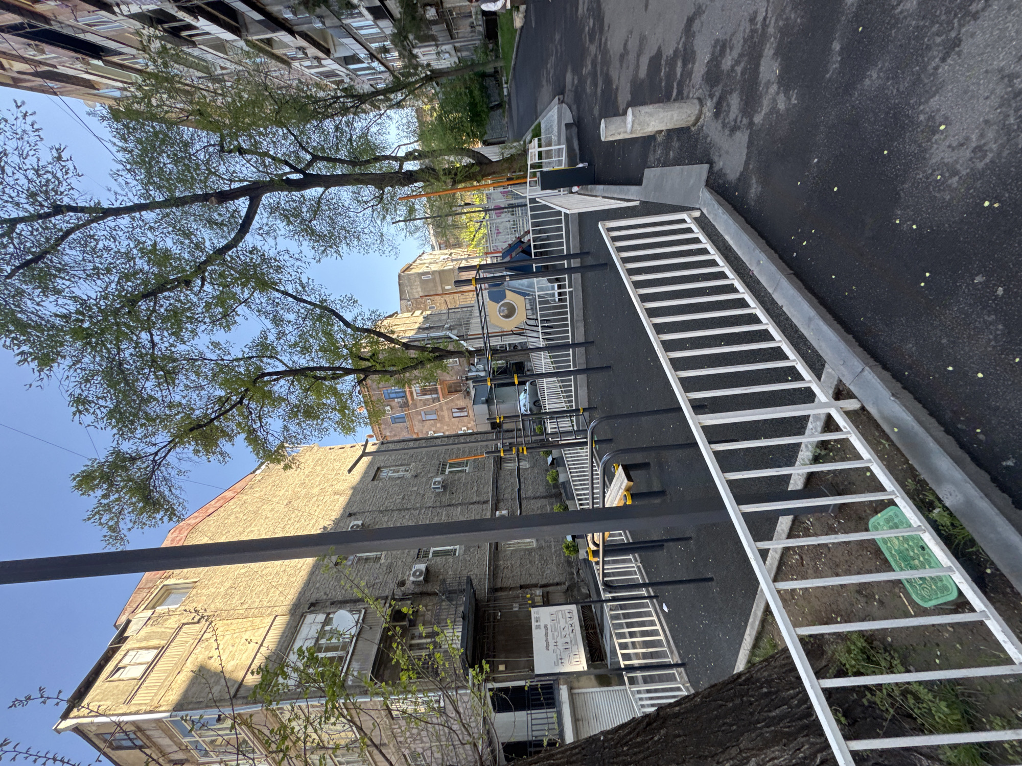 This image depicts an outdoor urban scene, likely a residential courtyard or public exercise area in Yerevan, Armenia, captured during bright daylight with a clear blue sky. The original image is oriented sideways, but when viewed upright, it shows multi-story apartment buildings surrounding a paved space.
Prominent in the foreground and midground is an exercise area featuring various metal equipment, including horizontal bars, monkey bars, and a white, ladder-like structure lying flat. The ground in this area is dark asphalt, and a white metal fence partially encloses the space. A single person is faintly visible in the background, near the exercise equipment.
The buildings surrounding the area are typical urban residential structures, varying in age and style. Many have multiple windows, balconies, and air conditioning units. Some buildings feature brick or textured facades, while one has a distinctive light-colored, textured exterior and a reddish roof. Green trees with vibrant new leaves are abundant, suggesting spring or early summer, and cast shadows across the scene, indicating either morning or afternoon. A large white sign with black text, likely Armenian, is affixed to one of the buildings, but the details are illegible. A green mat is also visible on the ground near the exercise equipment. The scene conveys a functional and lived-in urban environment.