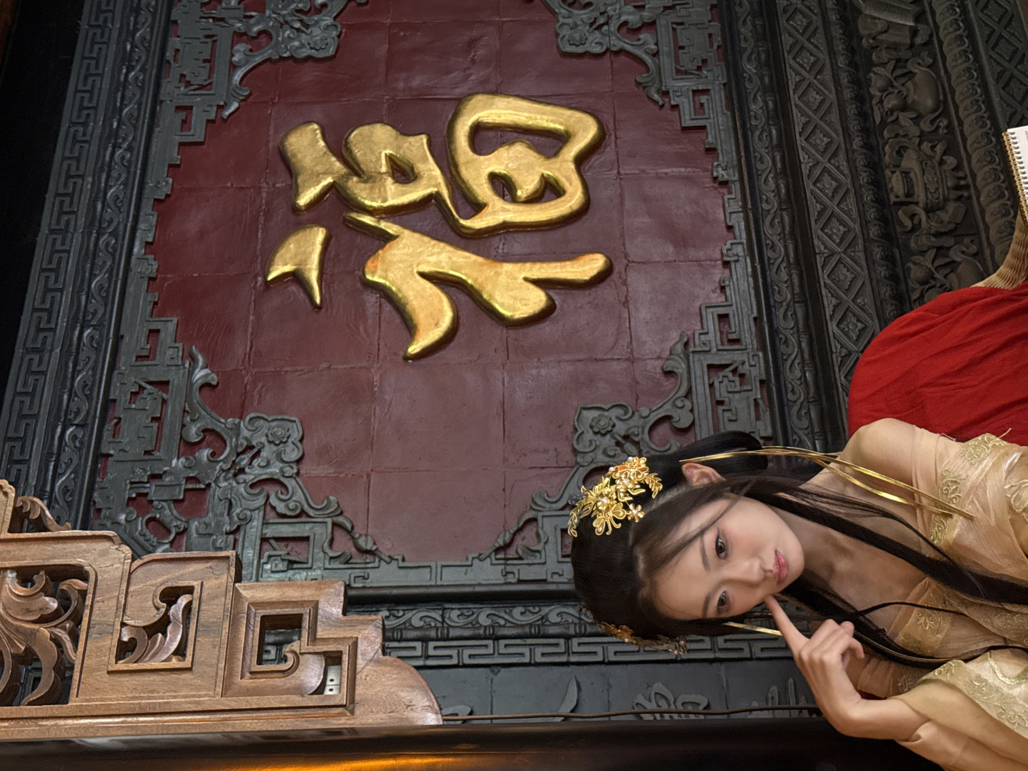 A woman with dark hair, adorned with gold hairpins, lies on a dark surface next to a wooden structure with intricate carvings. She wears a gold and beige dress with gold accents and is touching her face with one finger. Behind her is a red panel with golden Chinese characters, framed by ornate gray and black carvings. The setting is possibly Yuzhong District, China, suggested by the Chinese characters and architectural style.