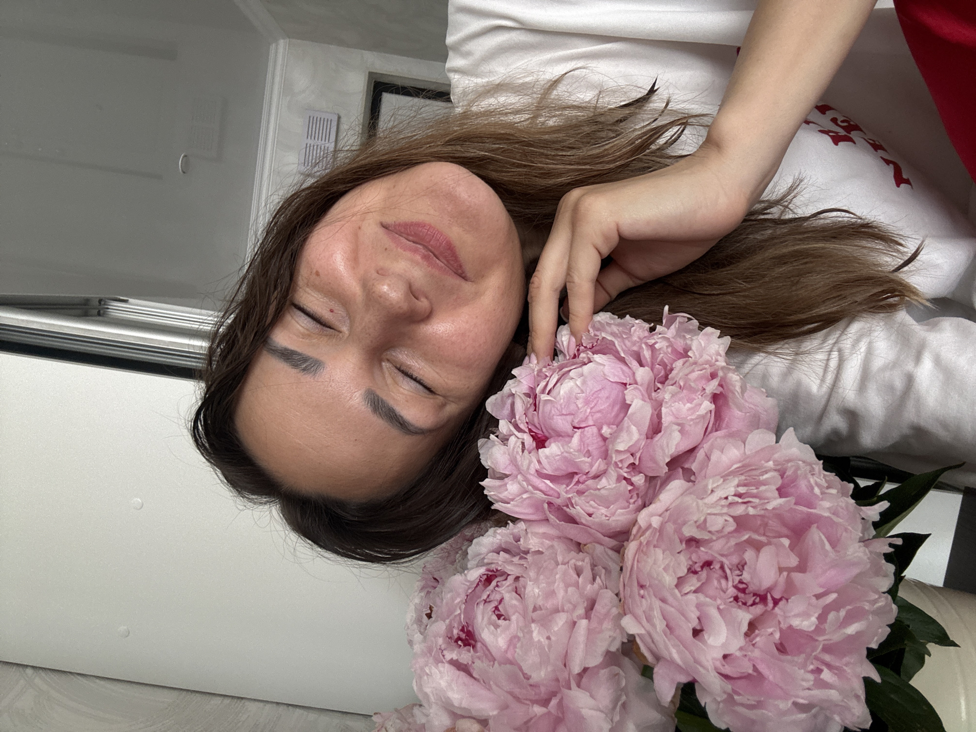 In a brightly lit indoor setting, a woman with closed eyes and a gentle smile rests her head against a bouquet of large, light pink peonies. Her hair is medium brown and falls loosely around her face and shoulders. She is wearing a white t-shirt. Her right hand is gently touching one of the peonies. The background is blurred but appears to be a domestic interior with white walls and a light-colored door or panel. There are no visible indications of the city of Sochi, Russia.
