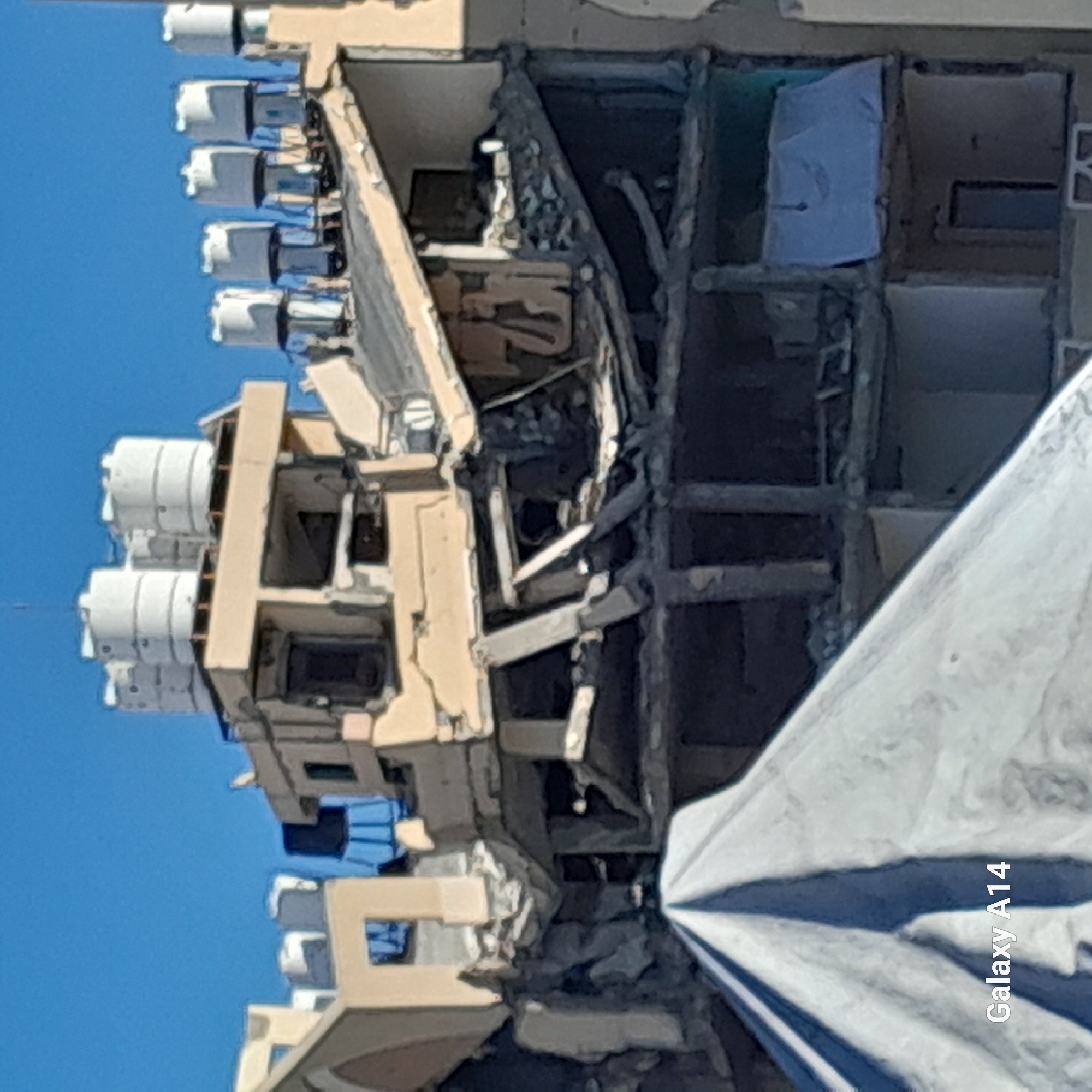 The image displays a multi-story building in Khan Yunis, Palestinian Territory, exhibiting severe structural damage. The upper levels are extensively compromised, with multiple floors partially collapsed and fractured reinforced concrete slabs and support beams prominently exposed. Interior spaces are visible through absent exterior walls, revealing internal debris and twisted structural components. The remaining rooftop sections support numerous white cylindrical water tanks, some appearing tilted due to the underlying damage. In the lower foreground, a large white tarp or tent structure partially obscures the building's lower stories. The background consists of a clear blue sky. No human figures are present within the frame.